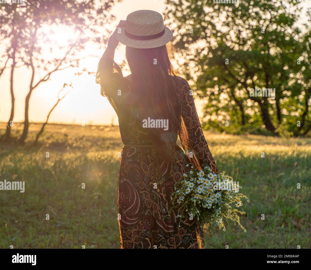 Eine glückliche Frau reist allein im Wald bei Sonnenuntergang, in einem langen Kleid, Hut und einem Blumenstrauß. Frohe Ferien. Rückansicht einer glücklichen Frau. Stockfoto