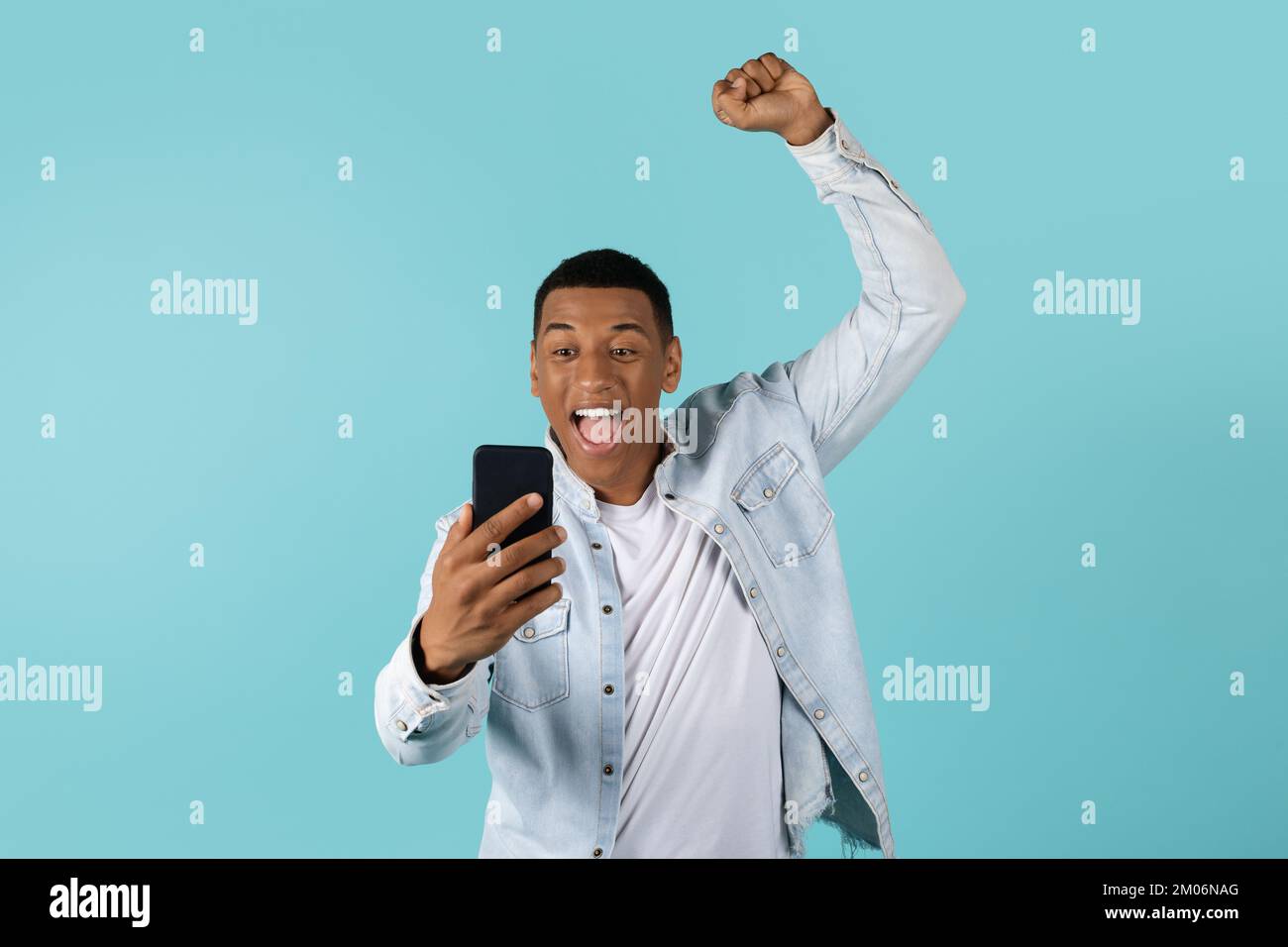 Freudig aufgeregter junger afroamerikanischer Mann in lässiger Atmosphäre mit offenem Mund Blick auf das Smartphone hebt die Hand Stockfoto