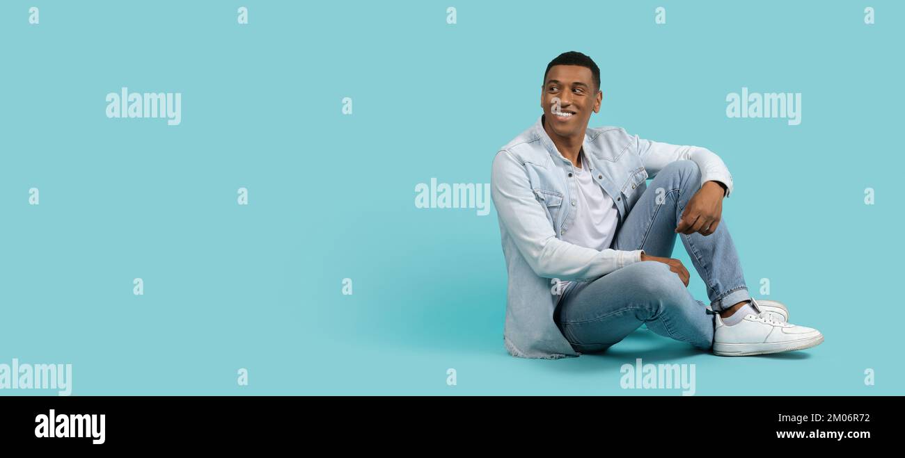 Lächelnder junger afroamerikanischer Mann in legerer Atmosphäre, auf dem Boden sitzen, auf freien Raum zurückblicken, Freizeit genießen Stockfoto
