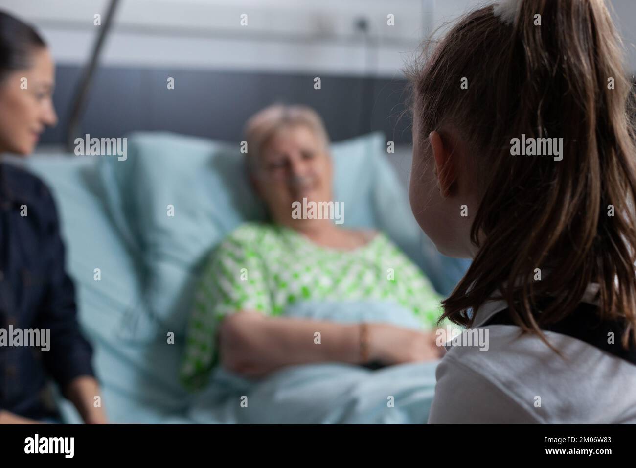 Ältere Frau im Fokus, lag im Krankenhausbett und unterhielt sich mit unidentifiziertem kleinen Mädchen. Familienbesuch bei einer genesenden älteren Patientin in der Klinik. Stockfoto
