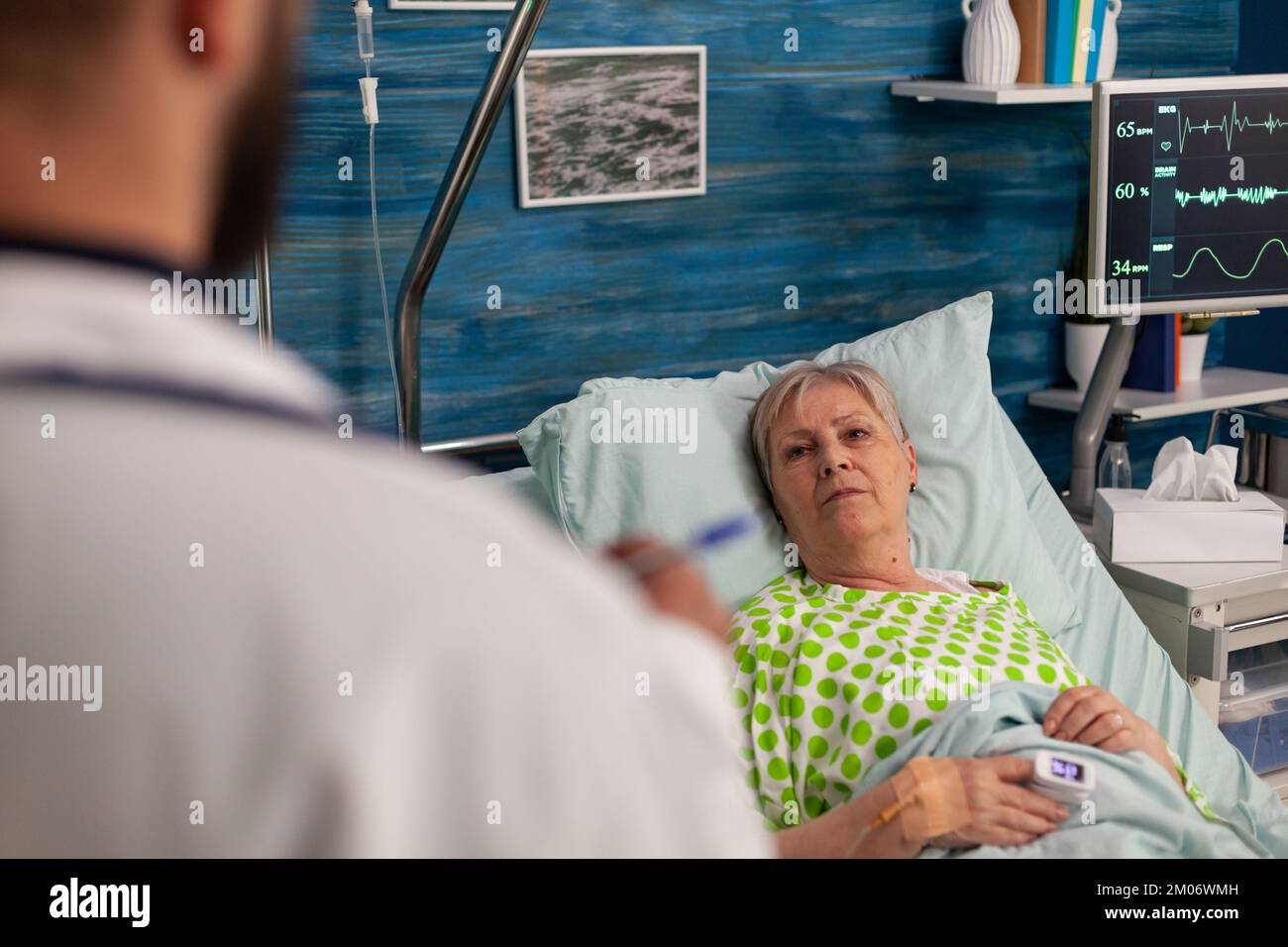 Ältere Frau, die im Krankenhausbett liegt und von einem männlichen Arzt betreut wird. Facharzt, der der kranken, hospitalisierten Patientin die Ergebnisse medizinischer Untersuchungen und Behandlungen erläutert. Stockfoto