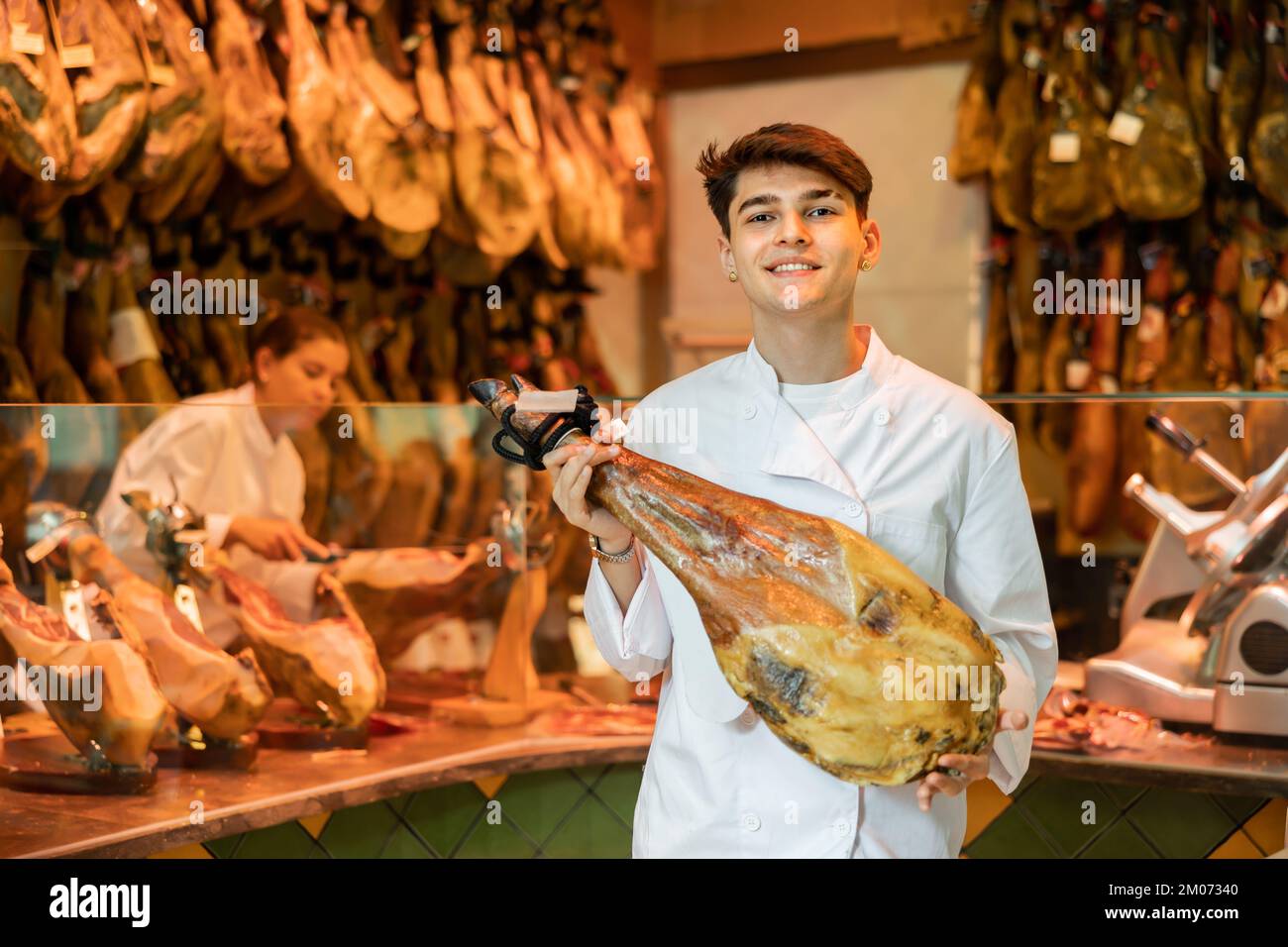 Fröhlicher junger Metzgerverkäufer, der ein Bein mit iberischem Schinken hält Stockfoto
