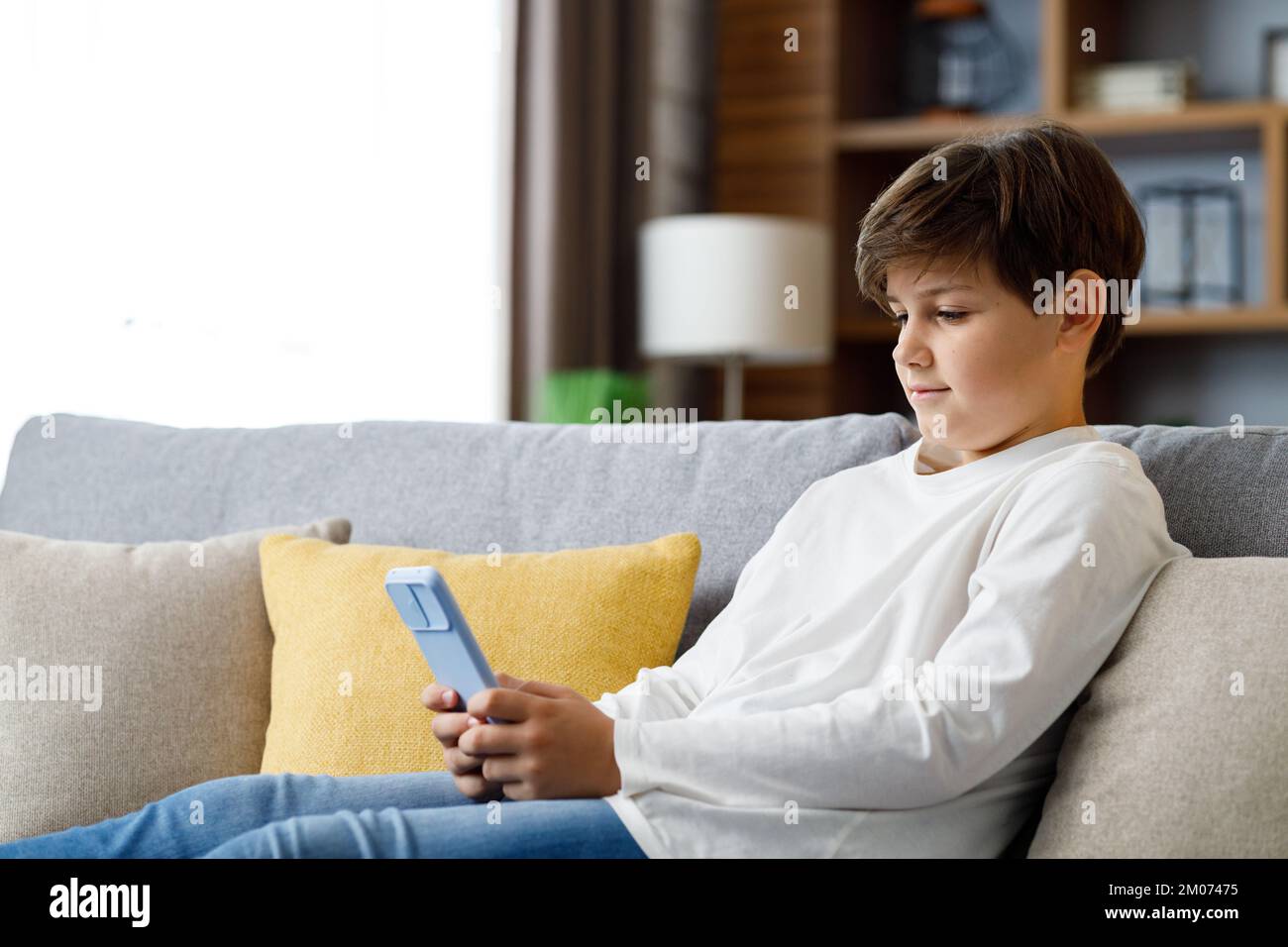 Portrait eines jungen niedlichen Jungen, der das Smartphone auf dem Sofa hält. Teenager zu Hause wählen Lieblingsmusik, SMS-Nachrichten, Surfen im Internet, beobachten Stockfoto