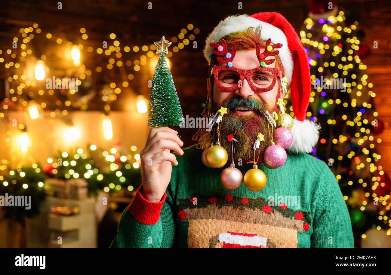 Weihnachtsmann in Partygläsern mit kleinem Weihnachtsbaum. Glücklicher Mann mit dekorativen Bällen im Bart. Stockfoto