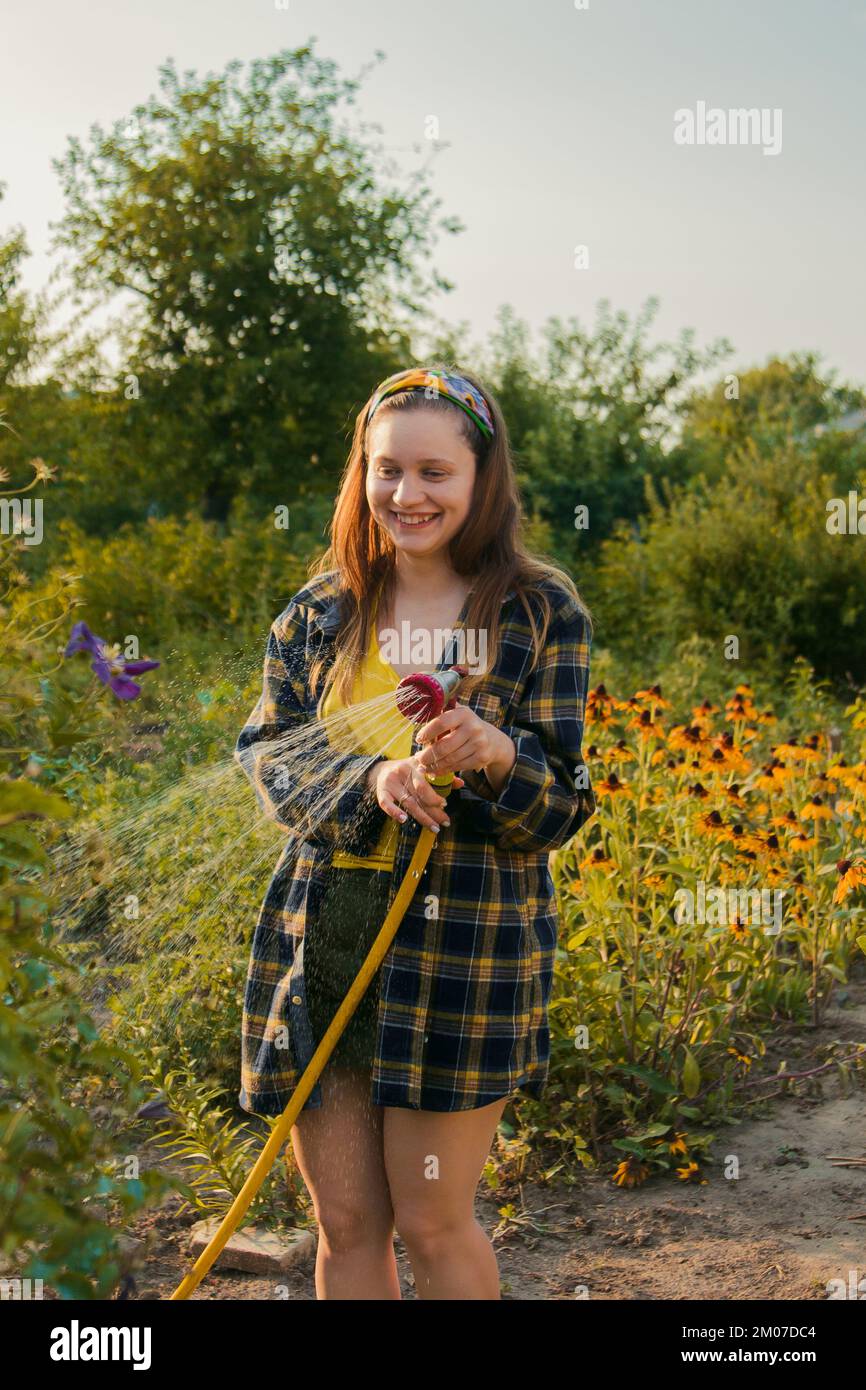 Ein hübsches Mädchen, das im Garten Spaß hat und Pflanzen mit einem Schlauch bewässert. Lächeln, während Sie ein Lieblingshobby nehmen Stockfoto