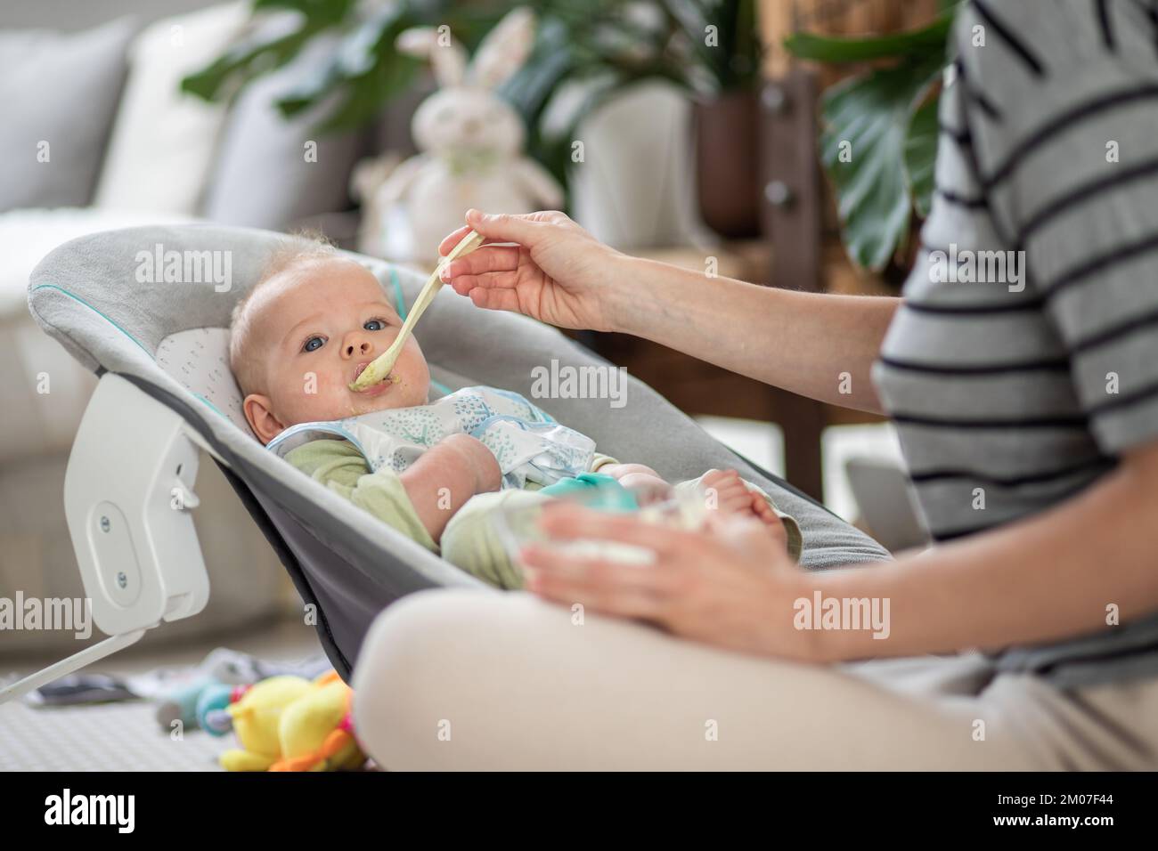 Mutter Löffel Fütterung ihres kleinen Jungen Kind in Babystuhl mit Fruchtpüree. Baby feste Nahrung Einführung Konzept Stockfoto