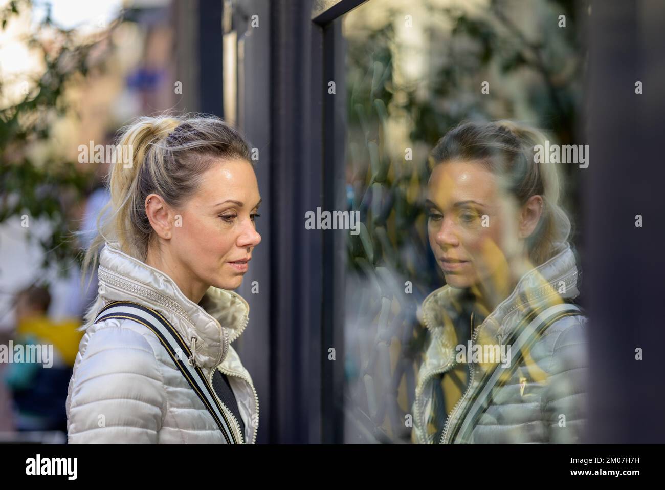 Seitenansicht einer sanft lächelnden, attraktiven Frau mittleren Alters mit blondem Haar und einem Pferdeschwanz, die interessiert durch das Fenster eines Shoppinggeschäfts blickt Stockfoto