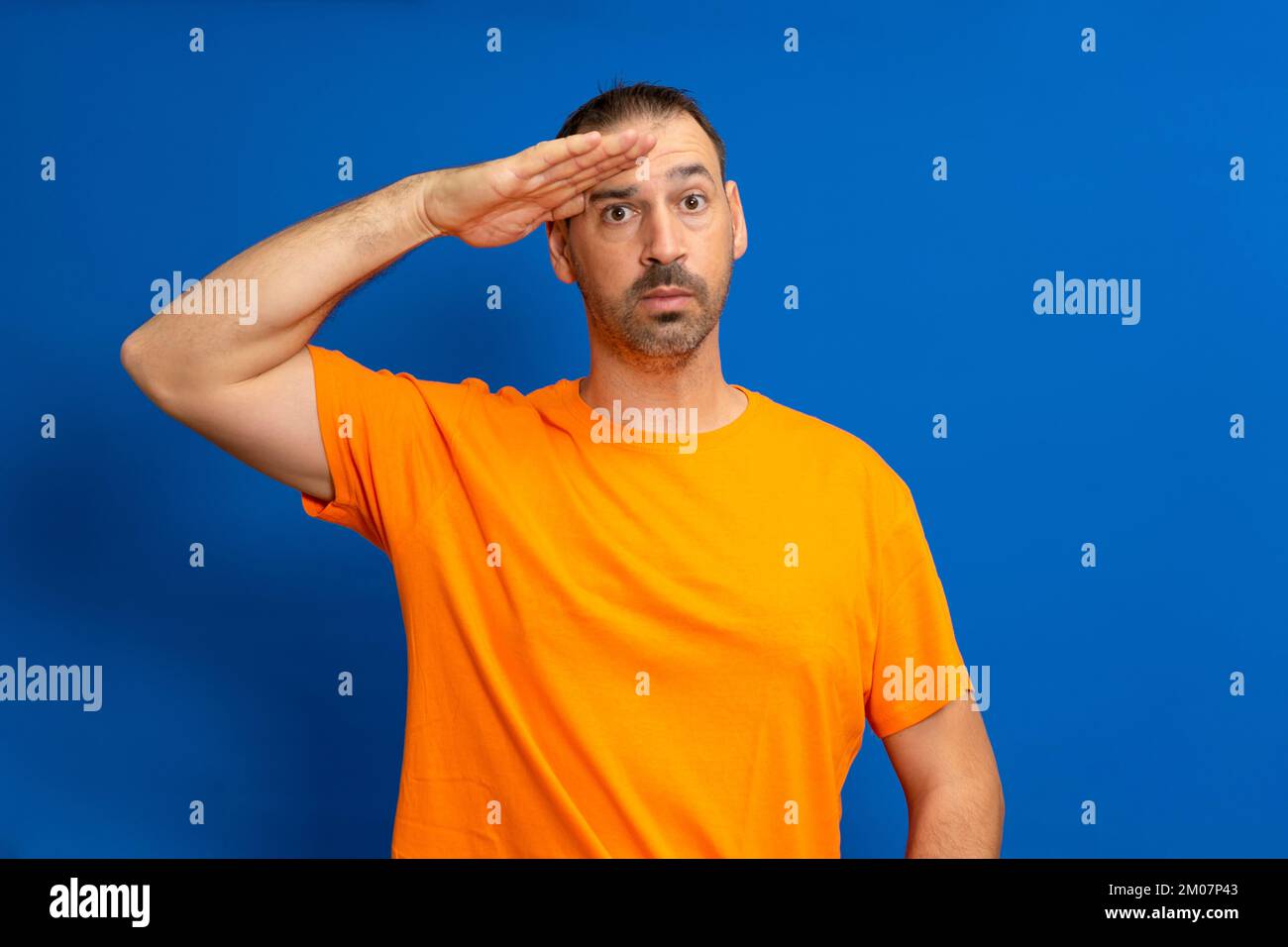 Ein Mann in Grußstellung und ernstem Gesicht. Ein lateinamerikanischer Mann mit einem Bart, der ein orangefarbenes T-Shirt trägt, isoliert auf blauem Hintergrund Stockfoto