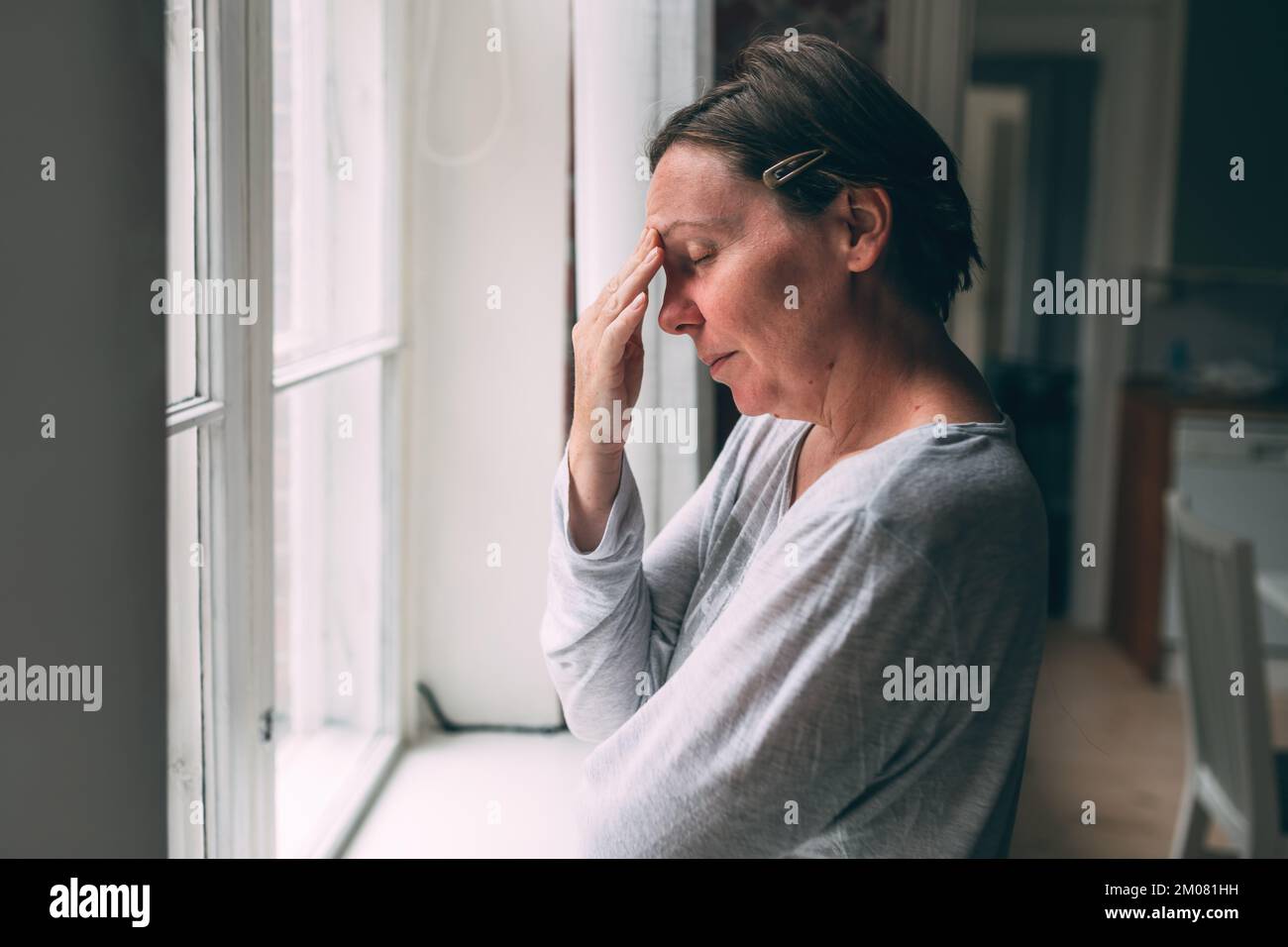 Reue und Reue, aufgebrachte mittelErwachsene weibliche Brünette, die morgens am Wohnzimmerfenster steht, selektiver Fokus Stockfoto