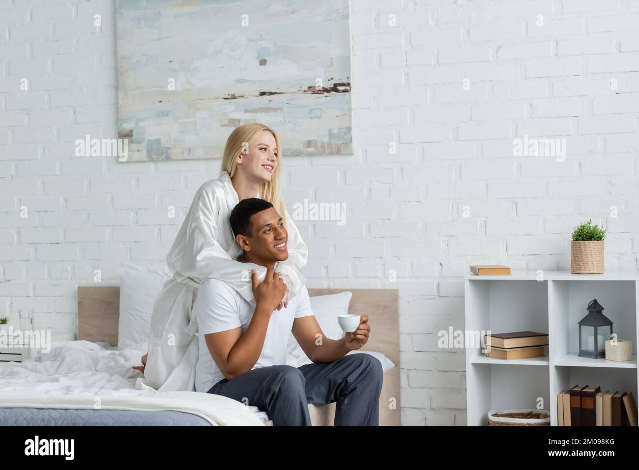 Junge Frau in weißem Seidenmantel umarmt einen afroamerikanischen Freund, sitzt mit Morgenkaffee im Bett Stockfoto