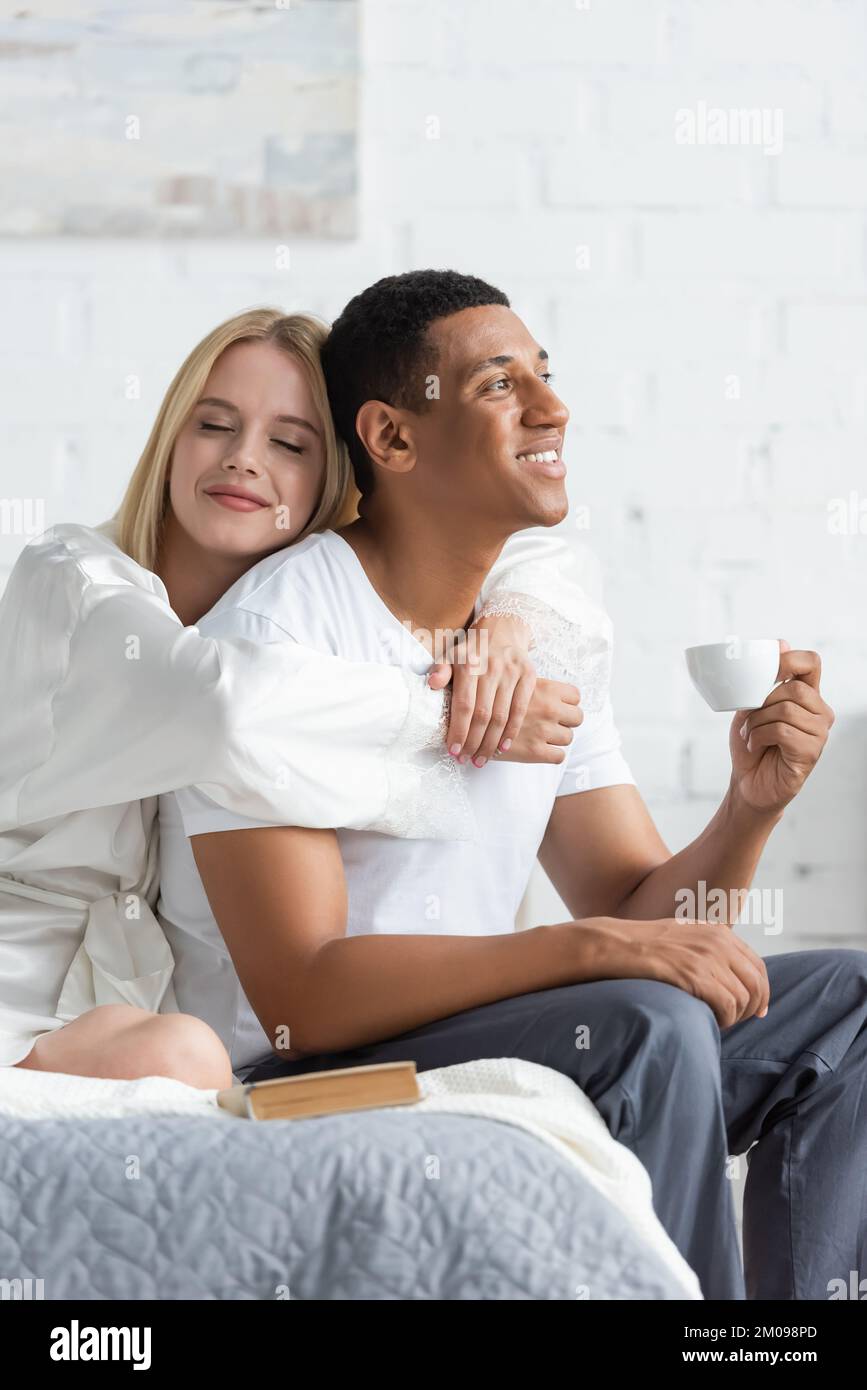 Junge blonde Frau mit geschlossenen Augen, die einen glücklichen afroamerikanischen Freund umarmt, der eine Kaffeetasse im Schlafzimmer hält Stockfoto