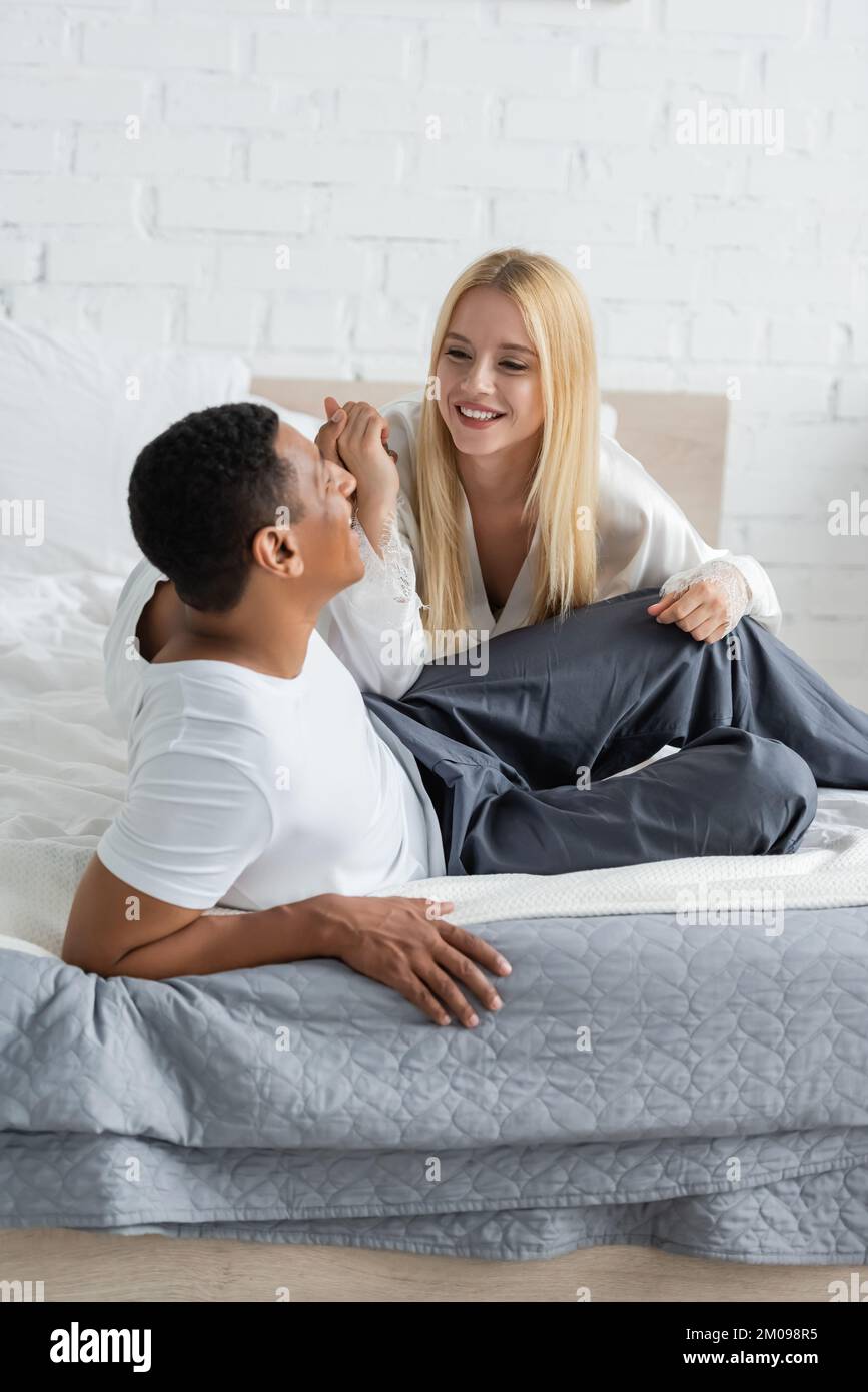Eine junge blonde Frau und ein afroamerikanischer Mann in Pyjamahosen, die Hände halten und sich im Bett anlächeln Stockfoto