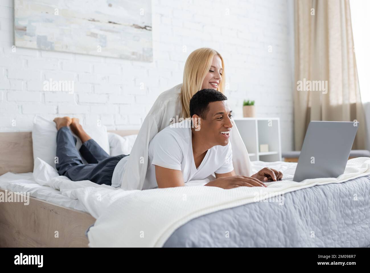 Junge blonde Frau lächelt neben einem fröhlichen afroamerikanischen Freund und sieht sich einen Film auf einem Laptop im Schlafzimmer an Stockfoto