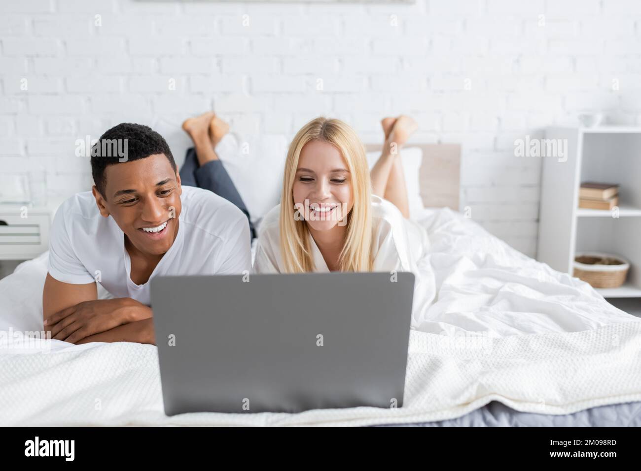 Blonde Frau mit afroamerikanischem Freund, die Comedy-Filme auf dem Laptop auf dem Bett sieht Stockfoto