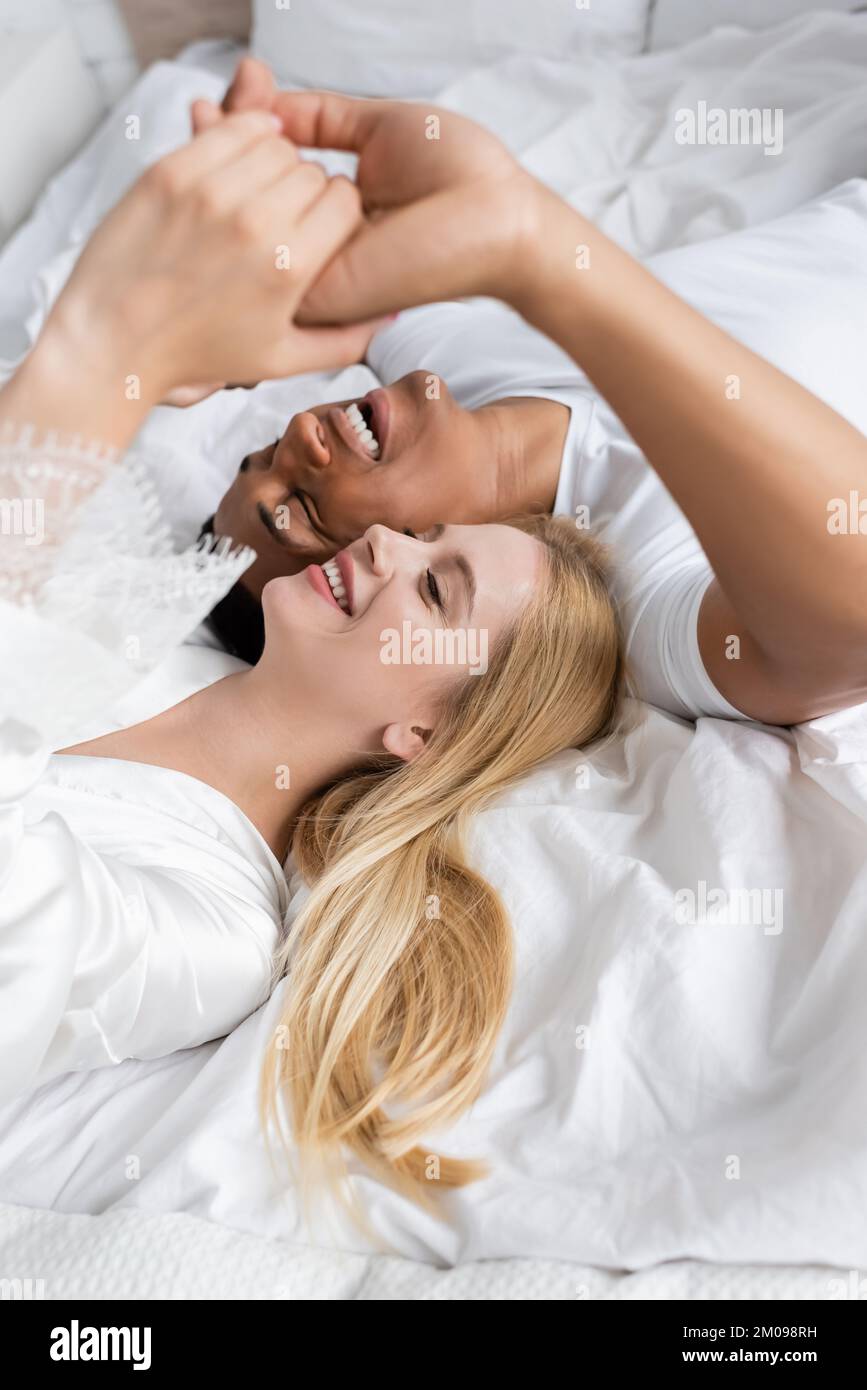 Blick von oben auf eine blonde Frau und einen afroamerikanischen Mann, der zu Hause Hände am Bett hält Stockfoto