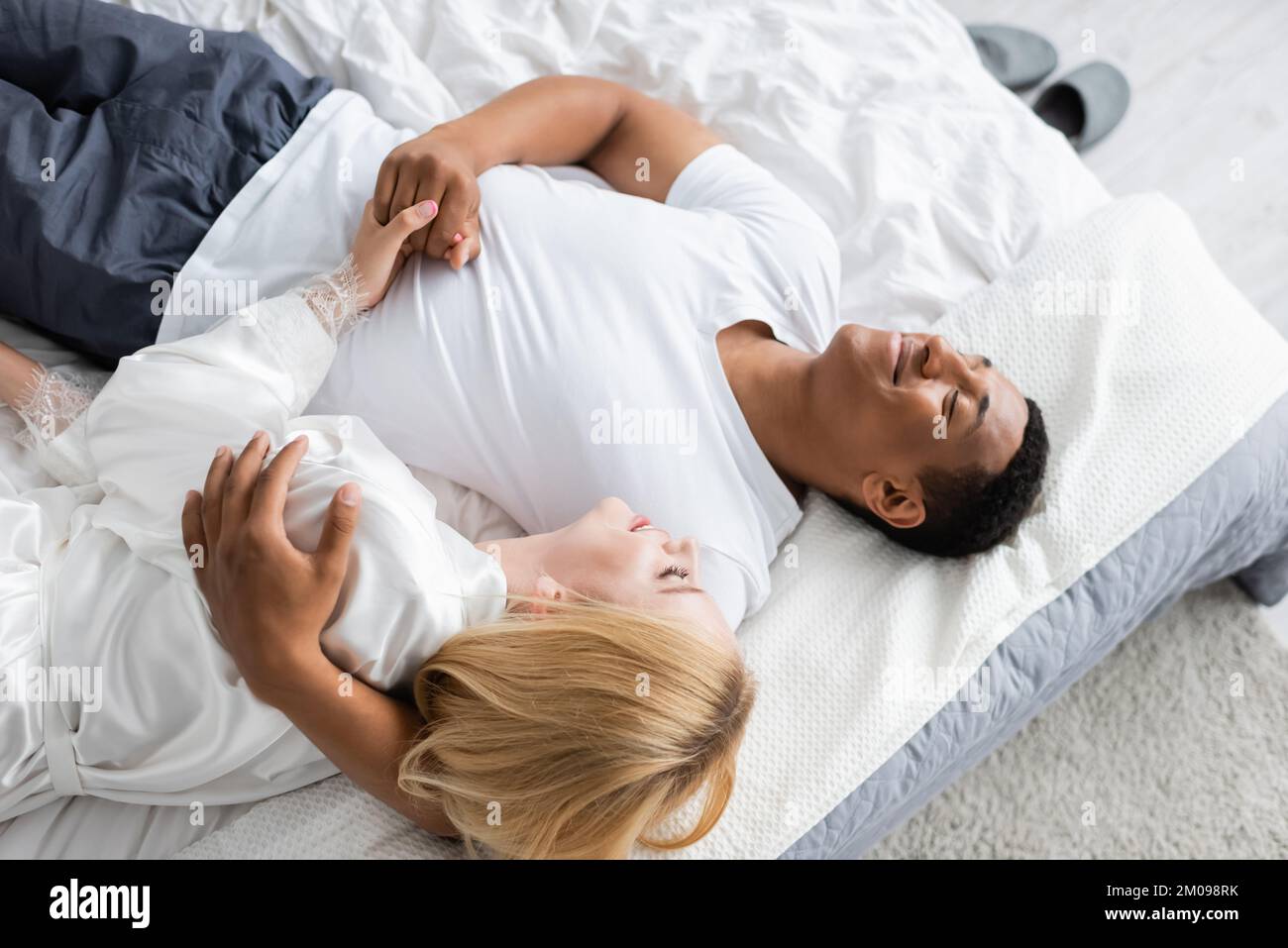 Ein afroamerikanischer Mann mit geschlossenen Augen, der eine blonde Frau umarmt und ihre Hand am Bett hält Stockfoto
