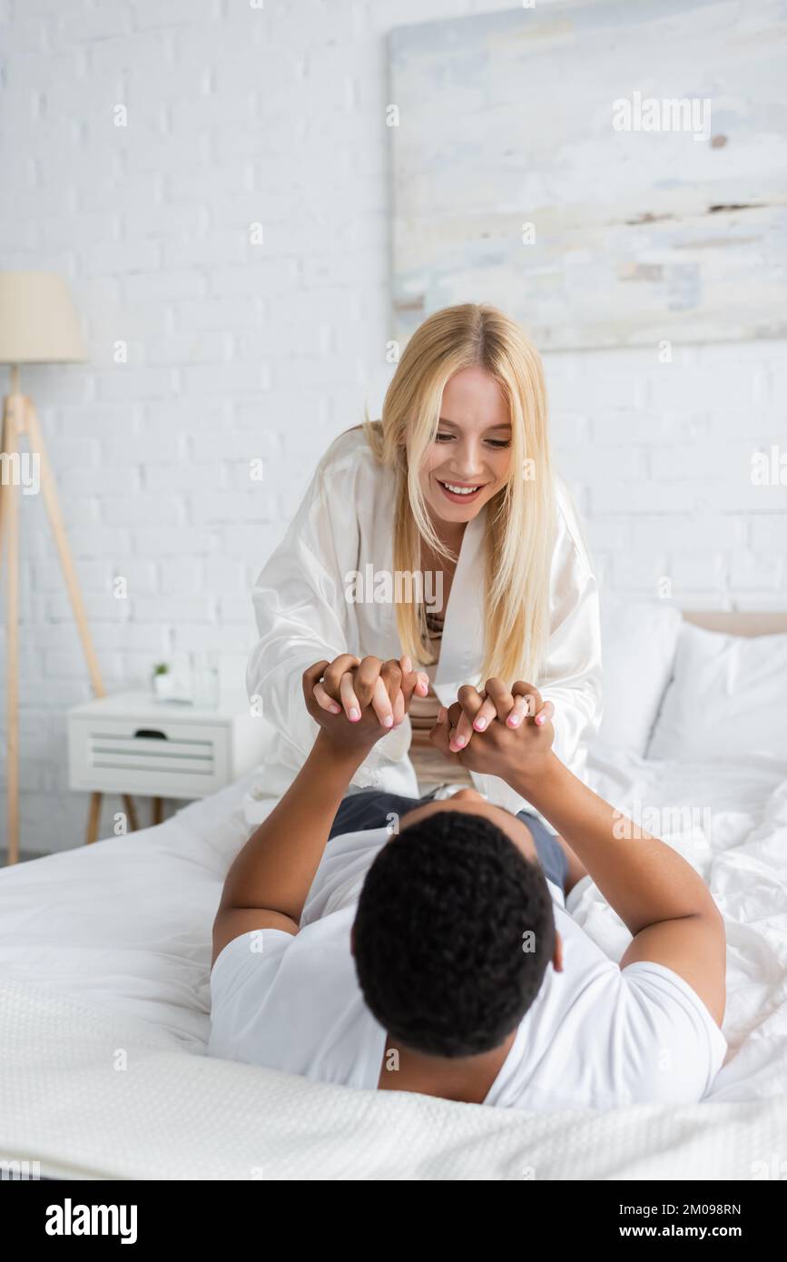 Aufgeregte blonde Frau, die Händchen eines afroamerikanischen Freundes hält, während sie Spaß im Schlafzimmer hat Stockfoto