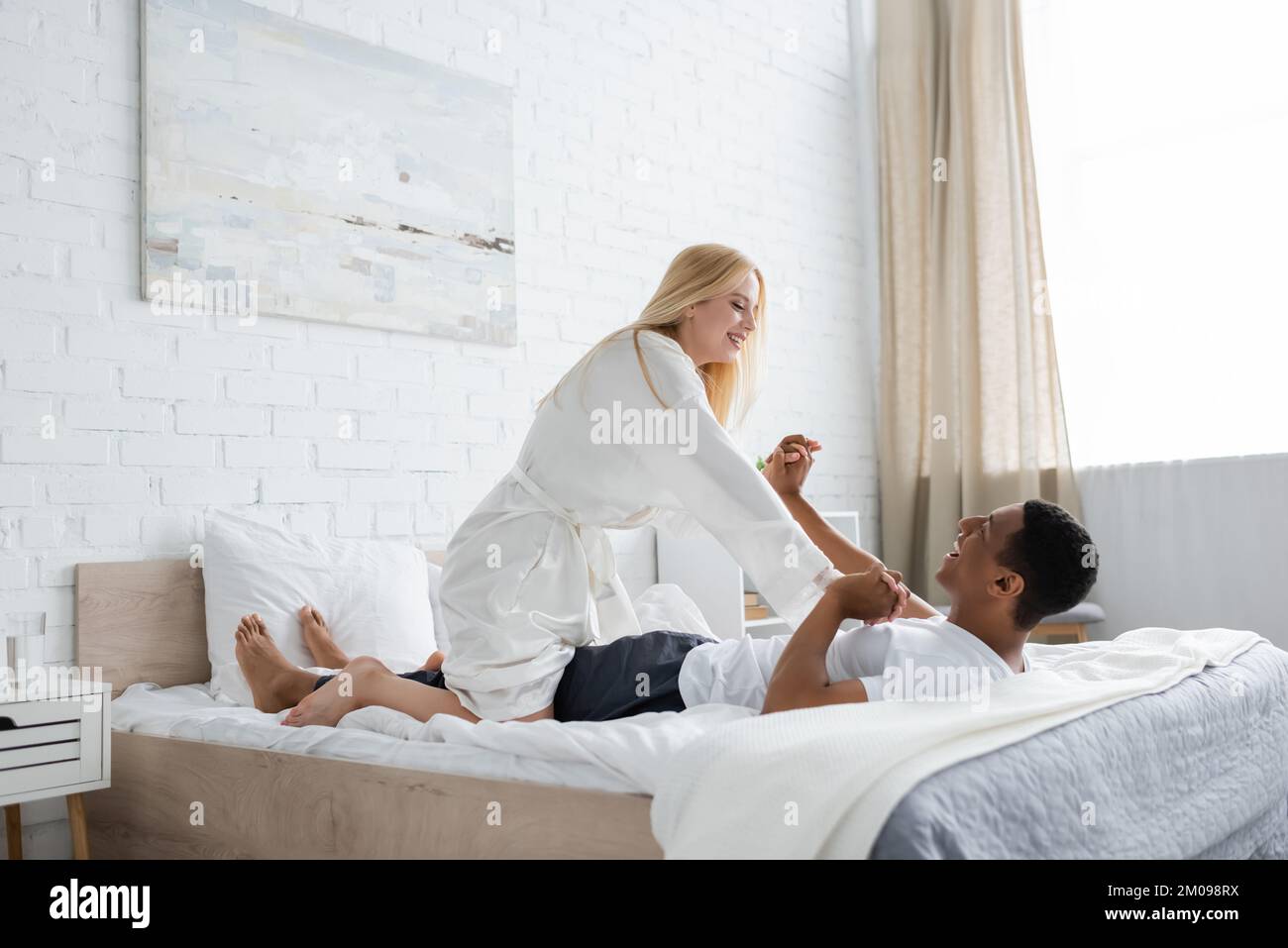 Fröhliche blonde Frau im weißen Bademantel, die Spaß mit einem afroamerikanischen Freund hat, der auf dem Bett liegt Stockfoto