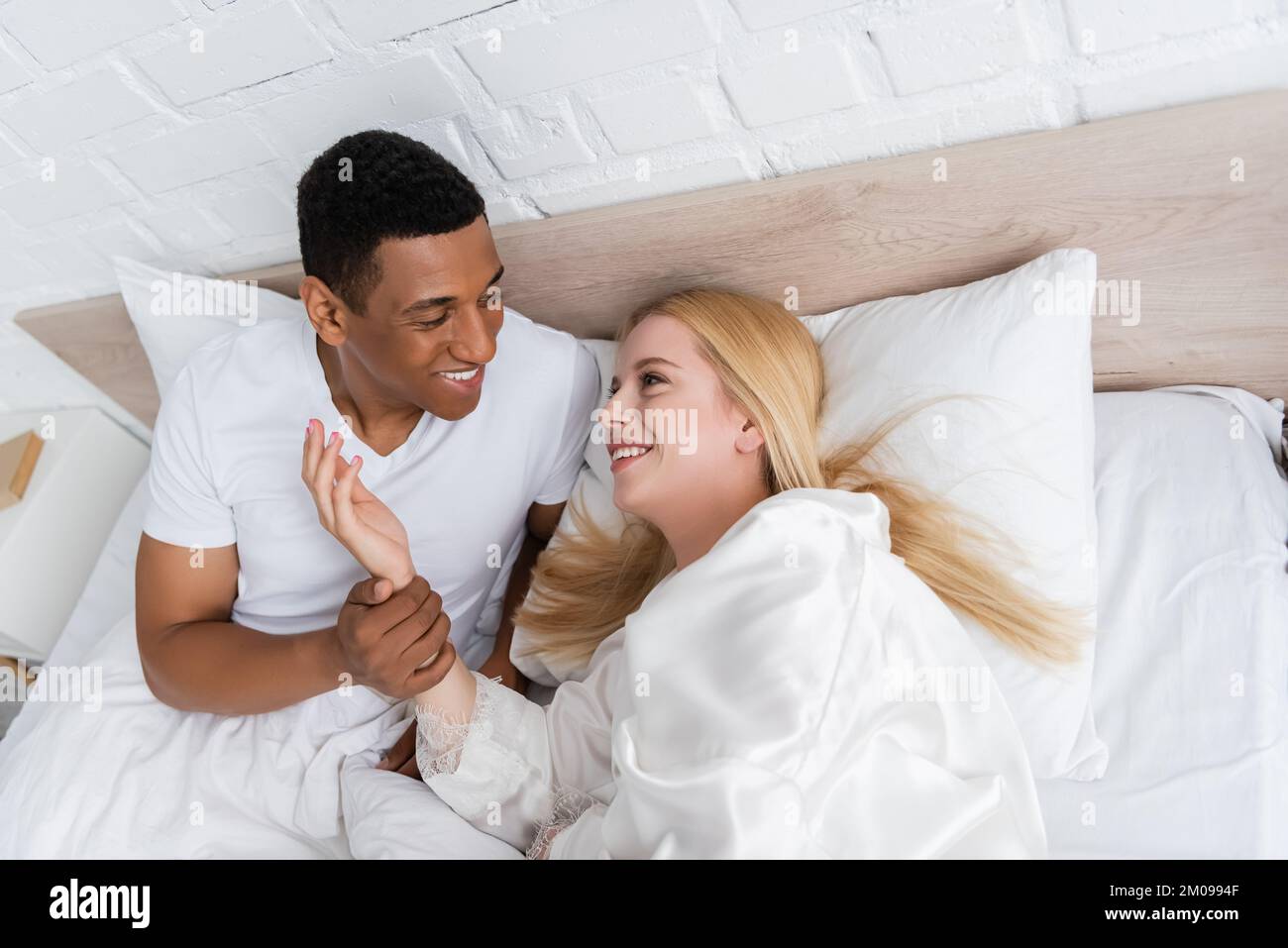 Aus einem hohen Blickwinkel kann man einen fröhlichen afroamerikanischen Mann sehen, der eine hübsche blonde Frau im Bett hält Stockfoto