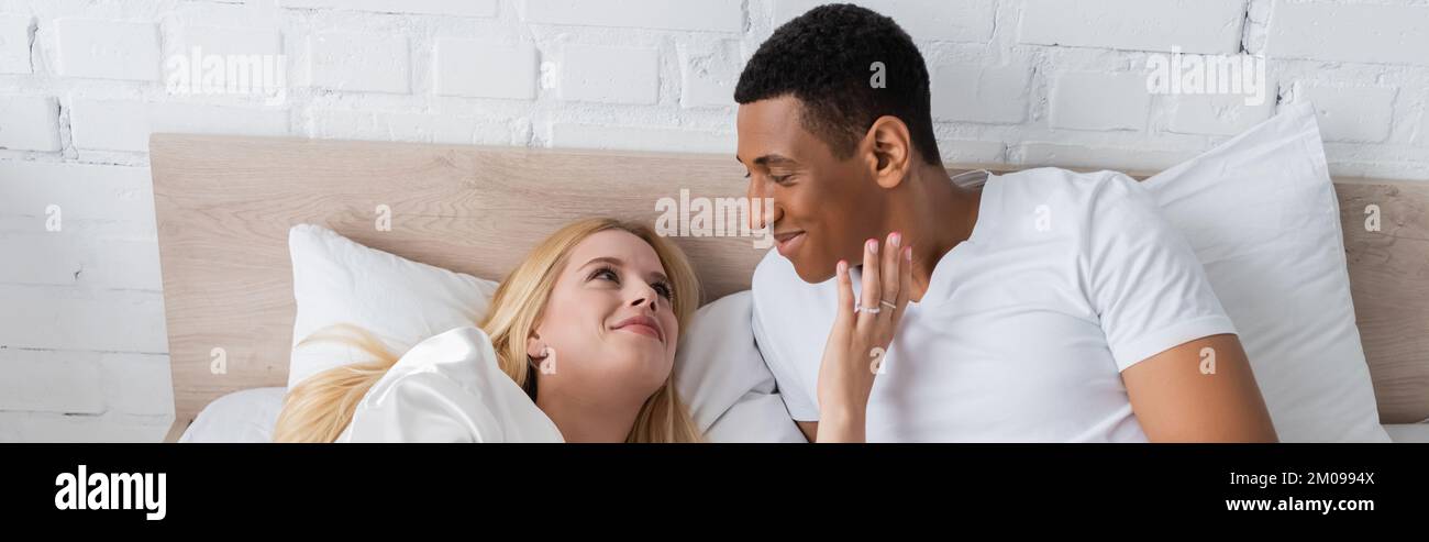 Lächelnde blonde Frau, die das Gesicht eines afroamerikanischen Freundes berührt, während sie morgens im Bett liegt, Banner Stockfoto