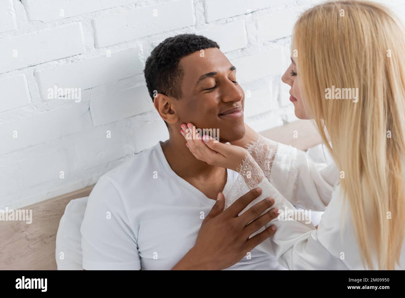 Junge blonde Frau, die das Gesicht eines afroamerikanischen Freundes berührt, der im Schlafzimmer mit geschlossenen Augen lächelt Stockfoto