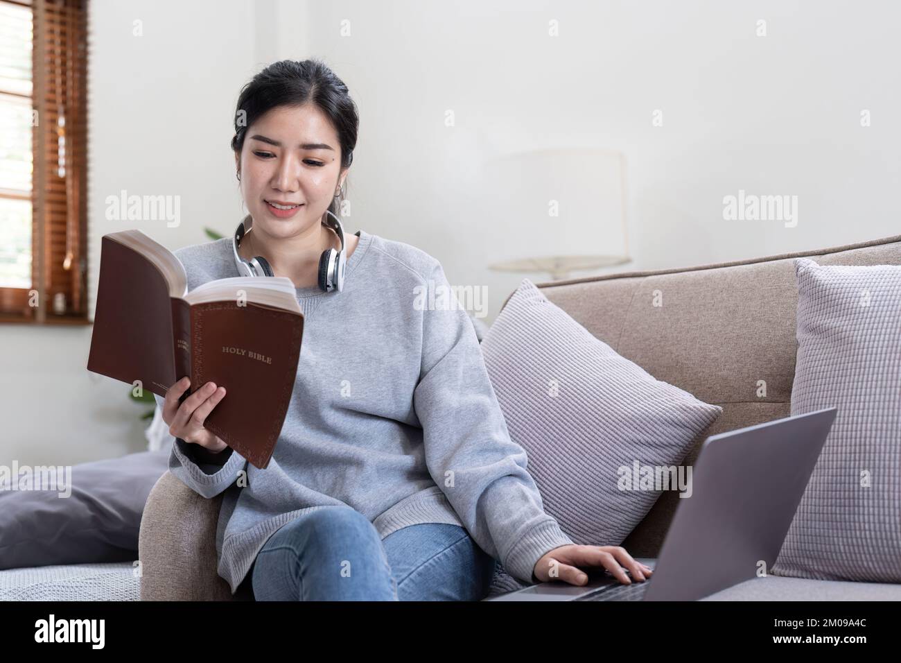 Junge, wunderschöne asiatische Studentin, die auf dem Sofa sitzt und zu Hause ein Buch liest, während sie ein Notebook für Online-Lernen verwendet Stockfoto