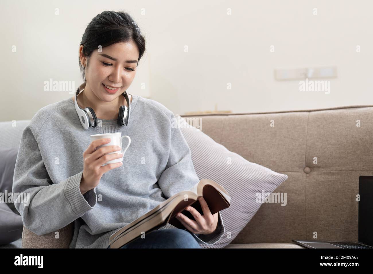 Junge, wunderschöne asiatische Studentin, die auf dem Sofa sitzt und zu Hause ein Buch liest, während sie ein Notebook für Online-Lernen verwendet Stockfoto