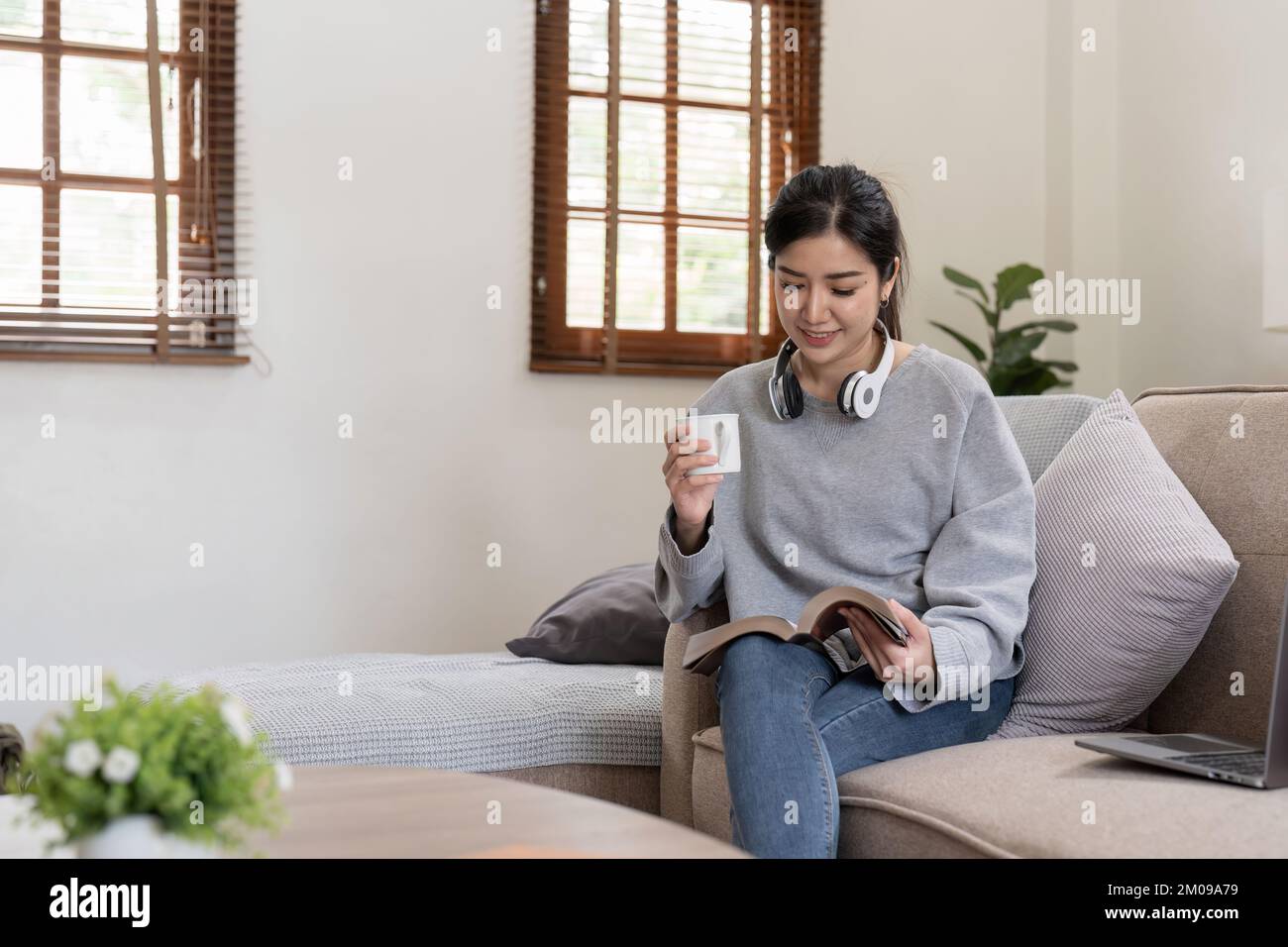 Junge, wunderschöne asiatische Studentin, die auf dem Sofa sitzt und zu Hause ein Buch liest, während sie ein Notebook für Online-Lernen verwendet. Stockfoto