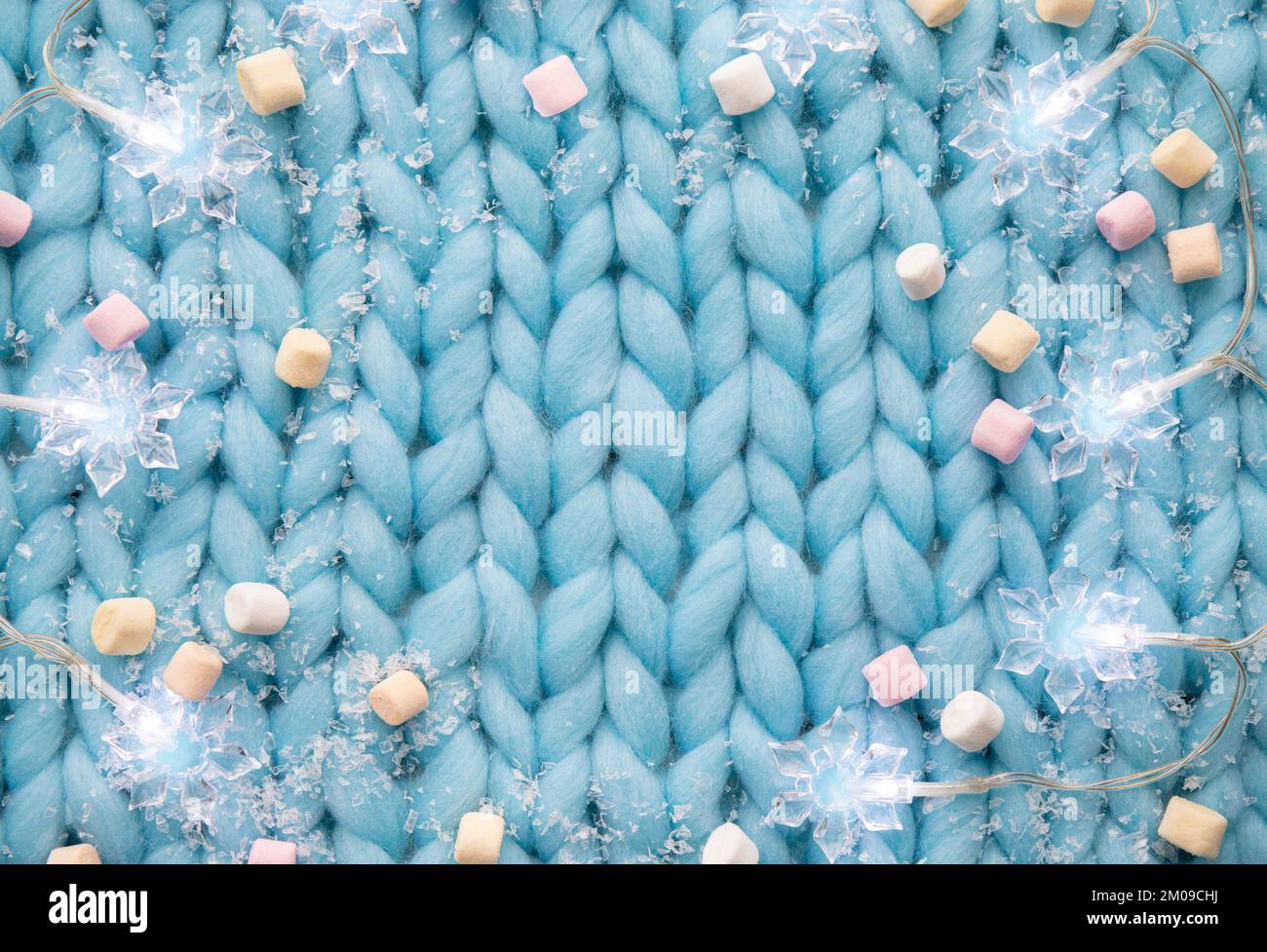 Blick über den hellblauen, weichen gestrickten Wollhintergrund mit Lichtern in Schneeflockenform, künstlichem Schnee und Mini-Marshmallows, Weihnachten Winter. Stockfoto