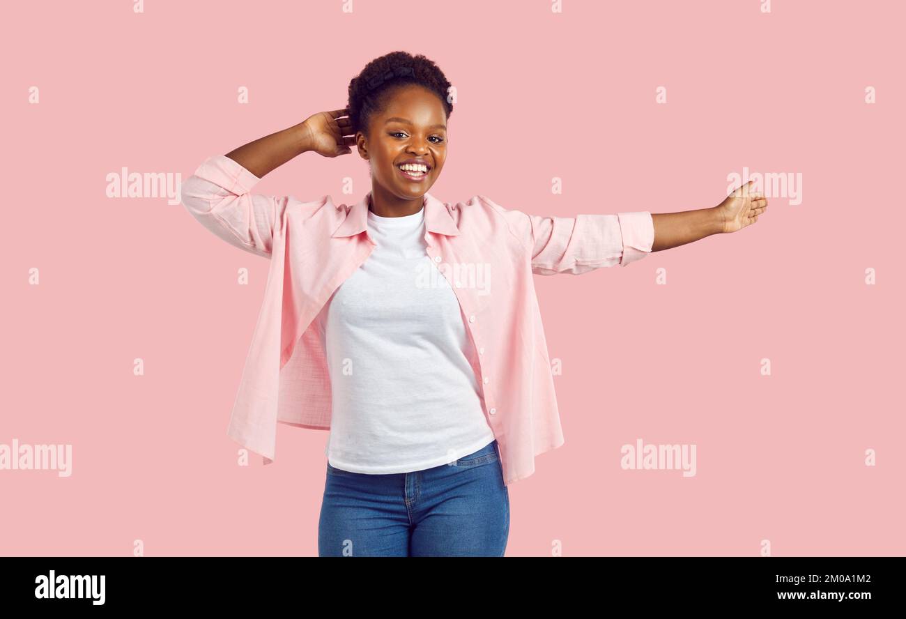 Fröhliche junge lockige, lachende afroamerikanische Frau in pinkem Hemd, Jeans, die auf pinkfarbenem Hintergrund tanzen. Stockfoto Fröhliche junge lockige, lachende afroamerikanische Frau in pinkem Hemd, Jeans, die auf pinkfarbenem Hintergrund tanzen. Stockfoto