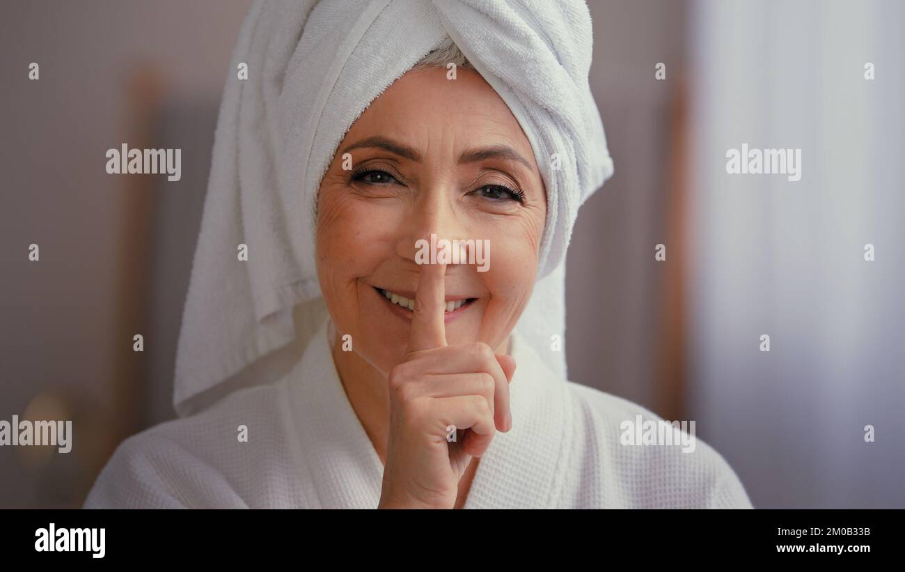 Alte, weiße, glückliche Frau, ältere Dame, weibliche Model mit Handtuch auf dem Kopf im Bademantel, die auf ein Lächeln der Kamera schaut und eine Geste der Stille zeigt Stockfoto