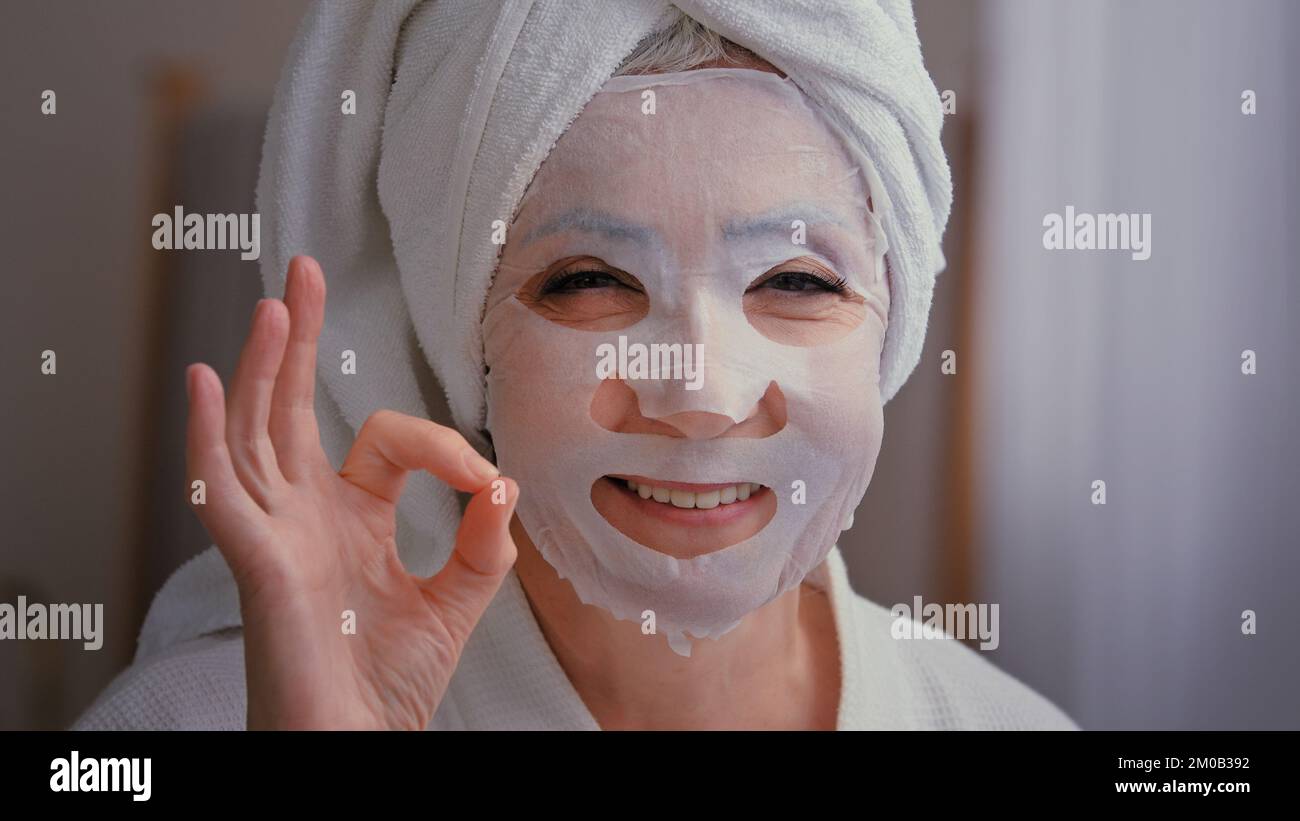 Nahaufnahme alte ältere ältere, reife weiße Frau mit Hautpflege Textil Hautmaske auf weiblichem Gesicht zeigt OK gute Schönheit Kosmetologie Verfahren 60s Lady Wrap Stockfoto