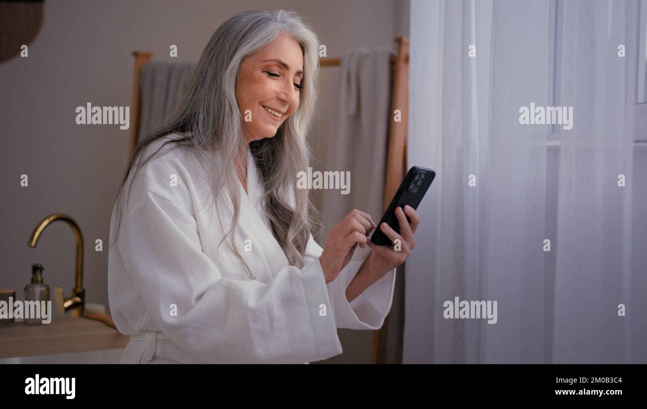 Seniorin, alte, weiße, reife Großmutter im Badezimmer im Bademantel, die auf die Telefonbuchung schaut und Kosmetikcreme zur Feuchtigkeitspflege bestellt Stockfoto