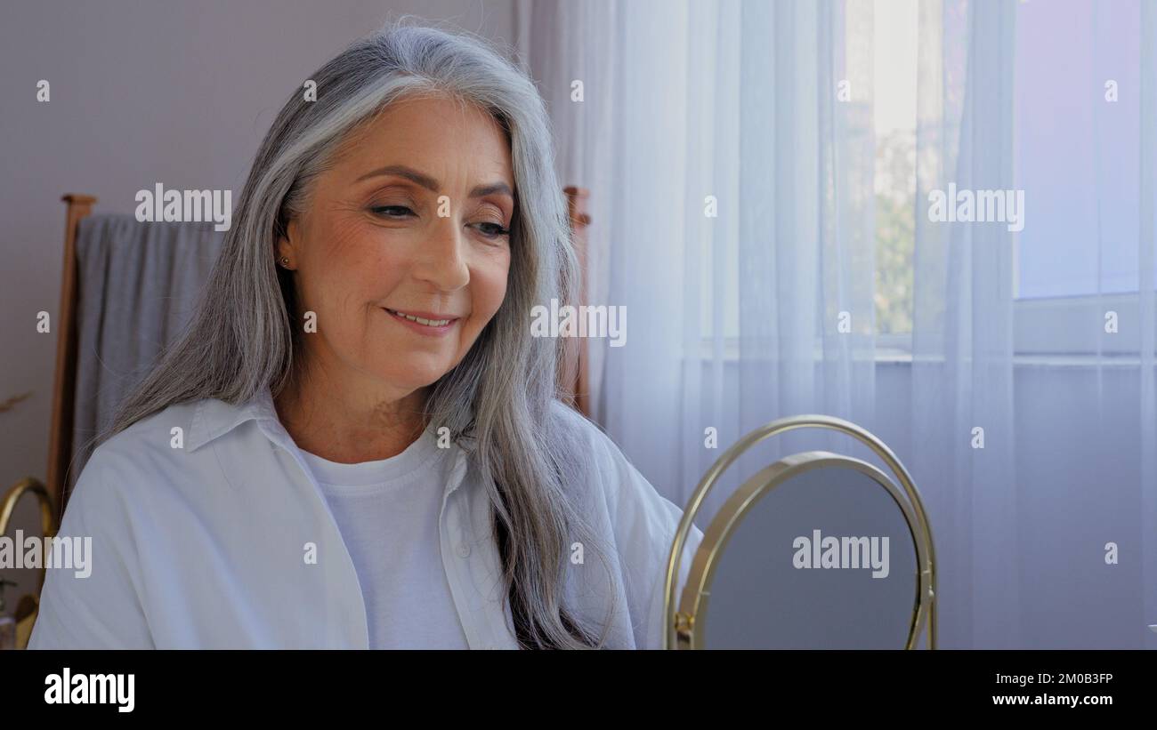 Kaukasische lächelnde, schöne alte ältere Dame, reife ältere Modellfrau mit grauen Haaren, weiblich, die auf den Spiegel schaut, lächeln, schau auf die Falte Stockfoto