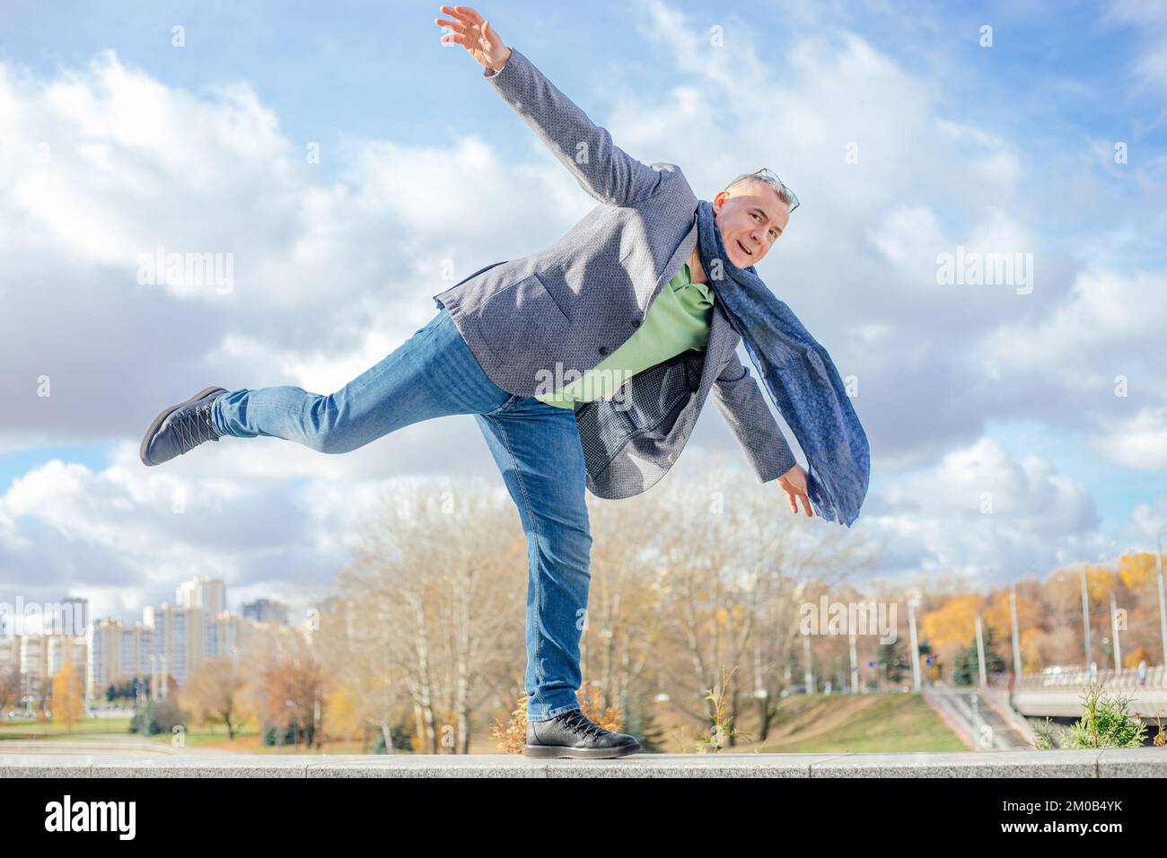 Porträt eines lustigen Mannes mittleren Alters mit kurzen Haaren, die auf einem Bein stehen, auf einer Betonbrücke stehen und sich die Hände strecken. Stockfoto