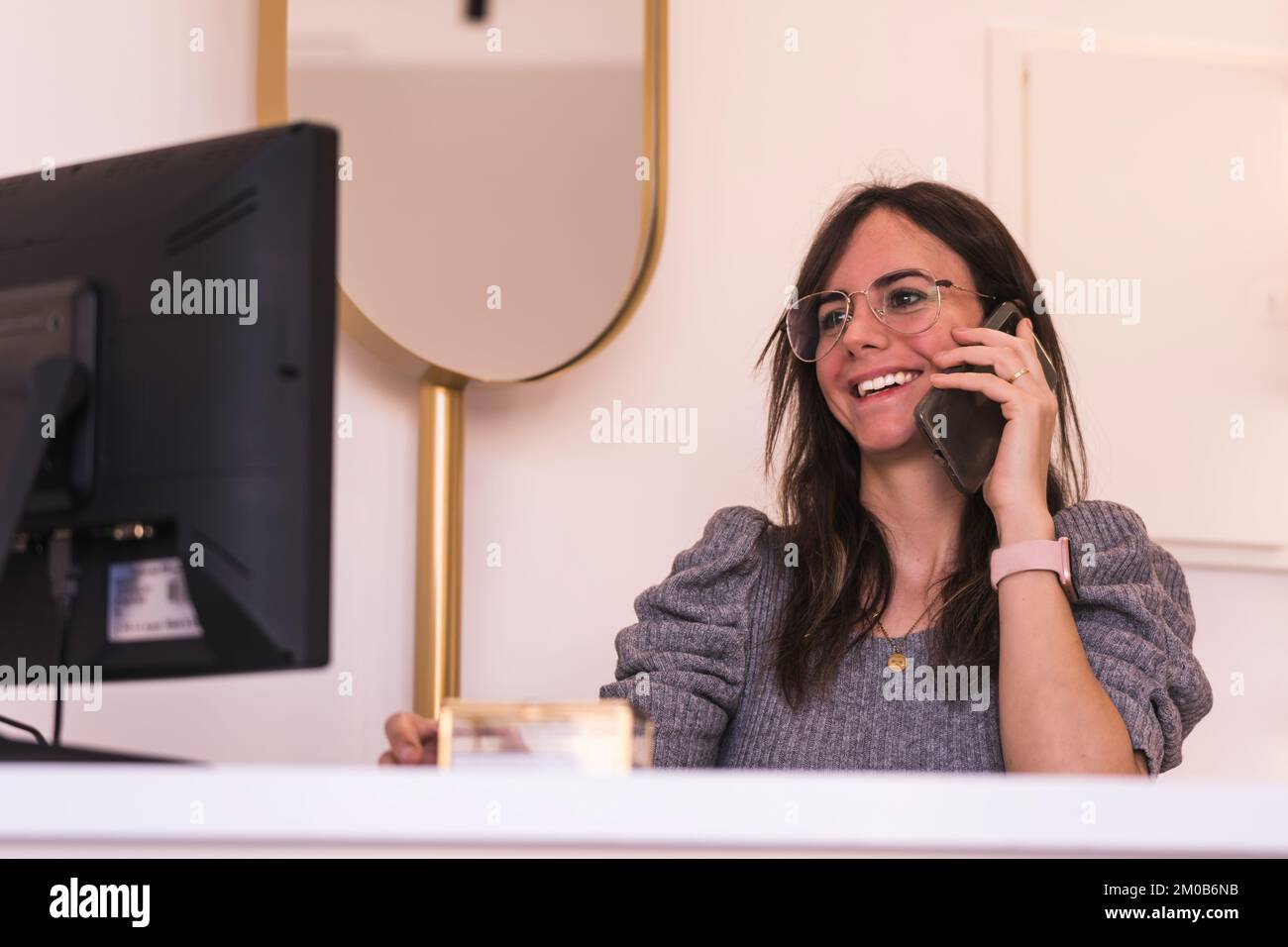 Brünette Rezeptionistin mit Brille lächelt, während sie ihren Computer benutzt und über ihr Smartphone spricht Stockfoto