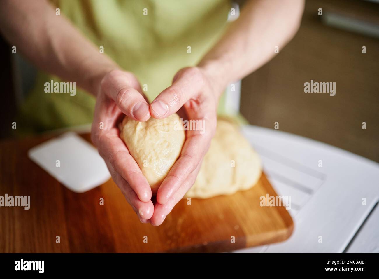 Arbeiten zu Hause Küchenkonzept, hausgemachtes Backen. Der Küchenchef in grüner Schürze hält die Teigkugeln in Händen, die in Herzform gefaltet sind. Hochwertiges Foto Stockfoto