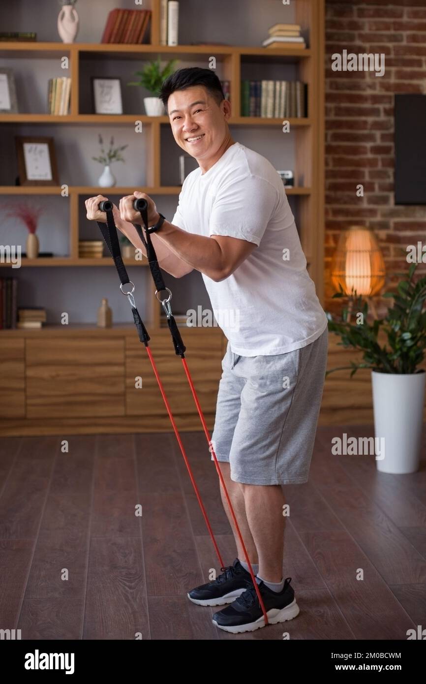 Aufgeregter, aktiver koreanischer Mann mittleren Alters, der mit einem Widerstandsband im Wohnzimmer trainiert und vor der Kamera lächelt Stockfoto