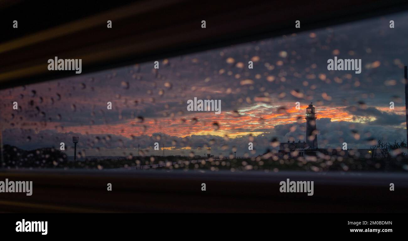 Blick auf den Sonnenaufgang von der Innenseite eines Wohnwagens an einem regnerischen Tag durch die Jalousien in Richtung Flamborough Head Leuchtturm und Leuchtfeuer auf der Landzunge Stockfoto
