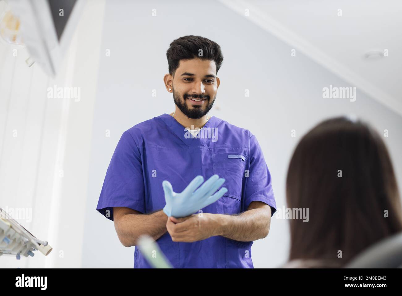 Ein selbstbewusster, bärtiger Zahnarzt trägt blaue Gummihandschuhe, steht in einer Zahnarztpraxis und ist bereit für die Untersuchung und Behandlung seiner Patientin. Glücklicher Patient und Zahnarztkonzept Stockfoto