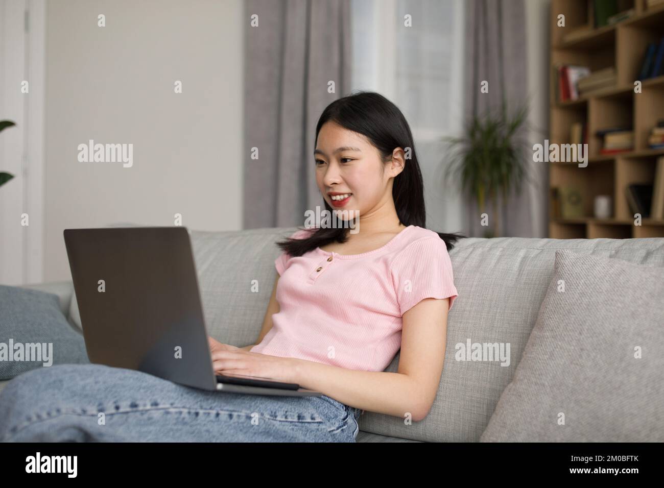 Fröhliche koreanische Freiberuflerin, die auf dem Sofa sitzt, die Laptop-Tastatur tippt und lächelt, Inneneinrichtung, kostenloser Kopierbereich Stockfoto