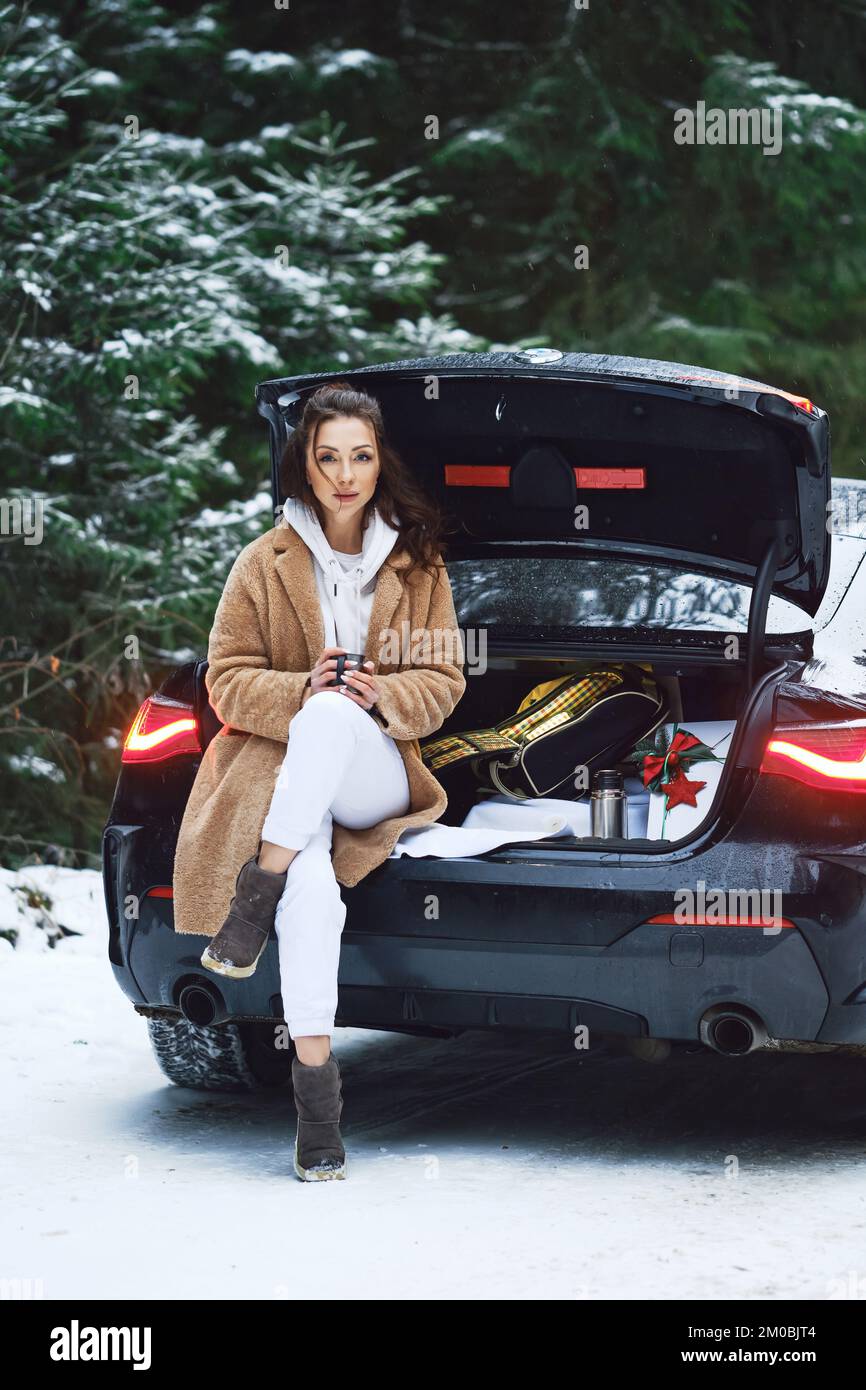 Frau sitzt auf einem Kofferraum mit Geschenkbox und trinkt heißen Kaffee auf der Winterstraße im Wald Stockfoto