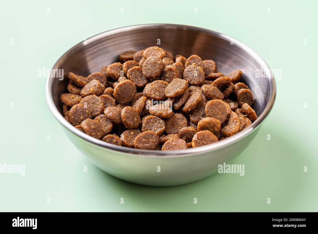 Trockenes Tierfutter in einer Metallschüssel auf grünem Hintergrund. Glutenfreie trockene Eiweißkibbles für Katzen in einer Edelstahl-Fütterschüssel. Vorderansicht. Stockfoto