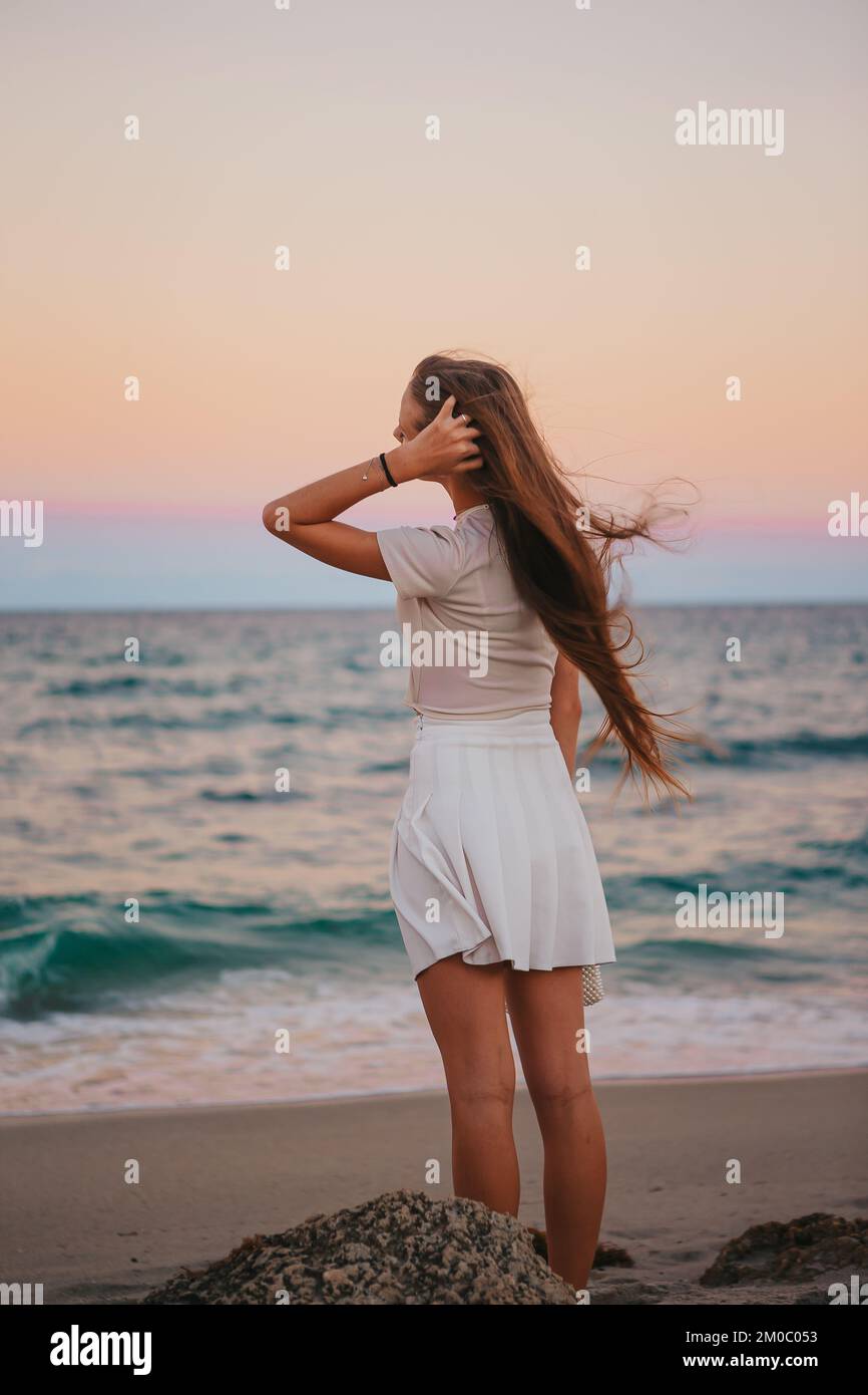 Bezauberndes, glückliches kleines Mädchen am Strand bei Sonnenuntergang mit Blick auf das Meer Stockfoto