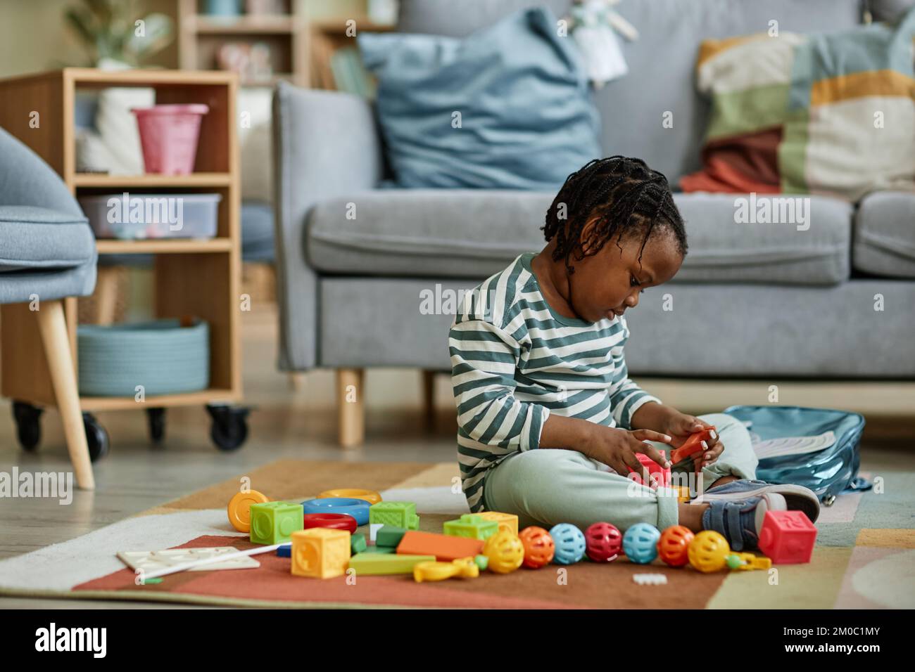 Seitliches Porträt eines süßen schwarzen Babys, das mit Spielzeug spielt, während es auf dem Boden in einem gemütlichen Zuhause sitzt, Kopierbereich Stockfoto