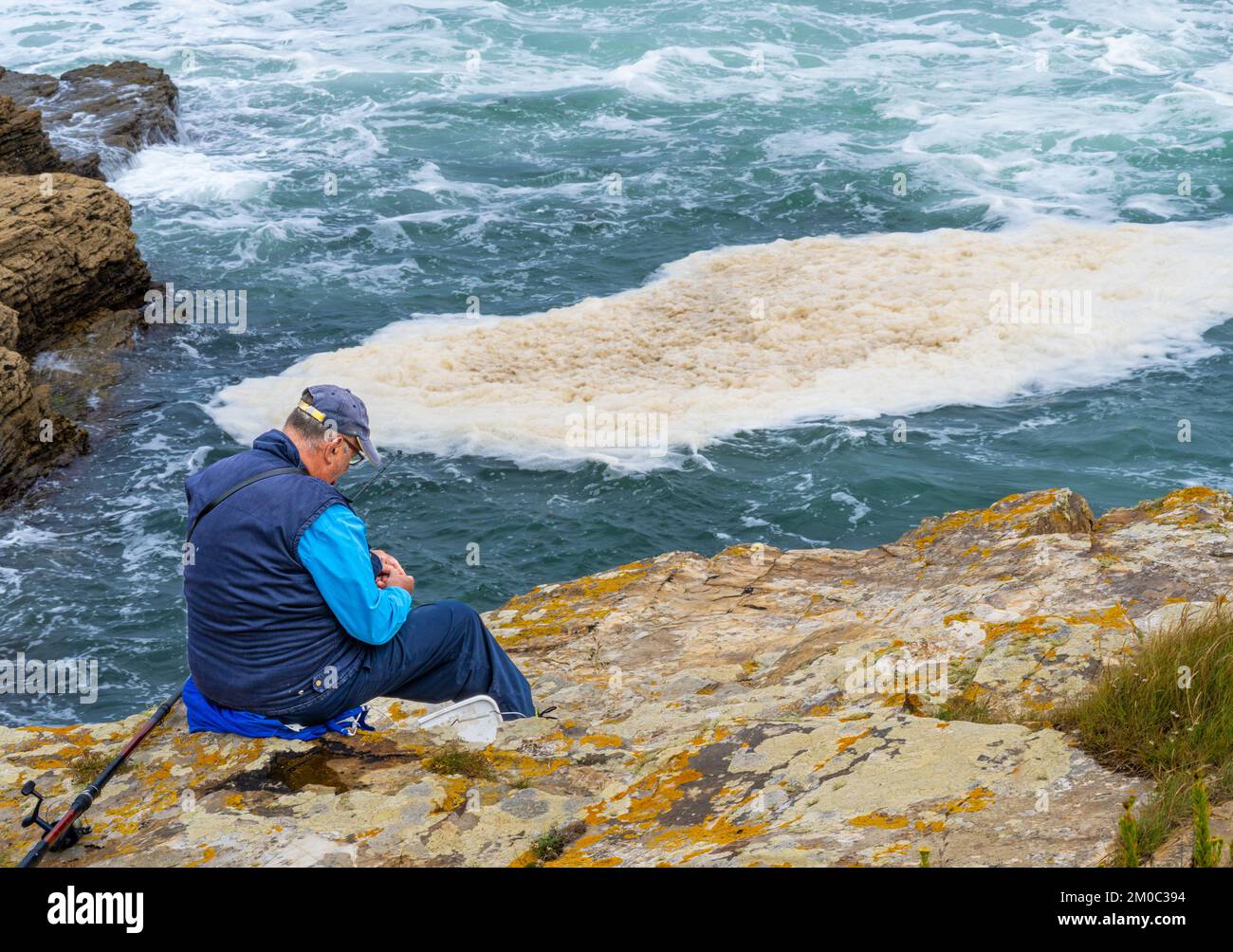 Ein älterer und pensionierter Mann, der seine Angelrute und Haken bereitete und auf den Klippen des Fischerdorfes Rinlo lag, mit dem Meer unter schlagenden Wellen. Stockfoto