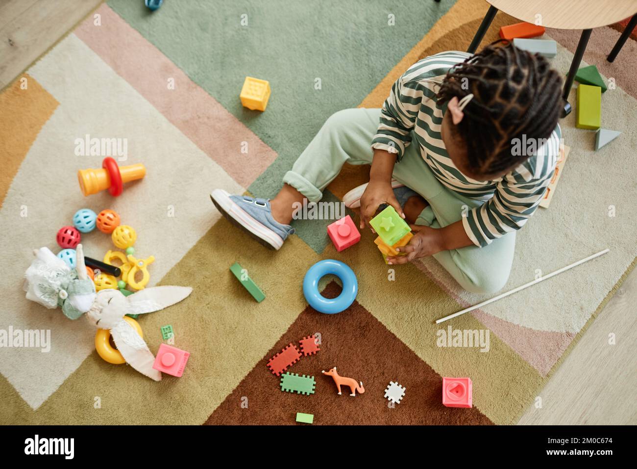 Blick von oben auf ein schwarzes Kleinkind, das zu Hause im Kinderzimmer mit bunten Spielzeugen auf einem gemütlichen Teppich spielt Stockfoto