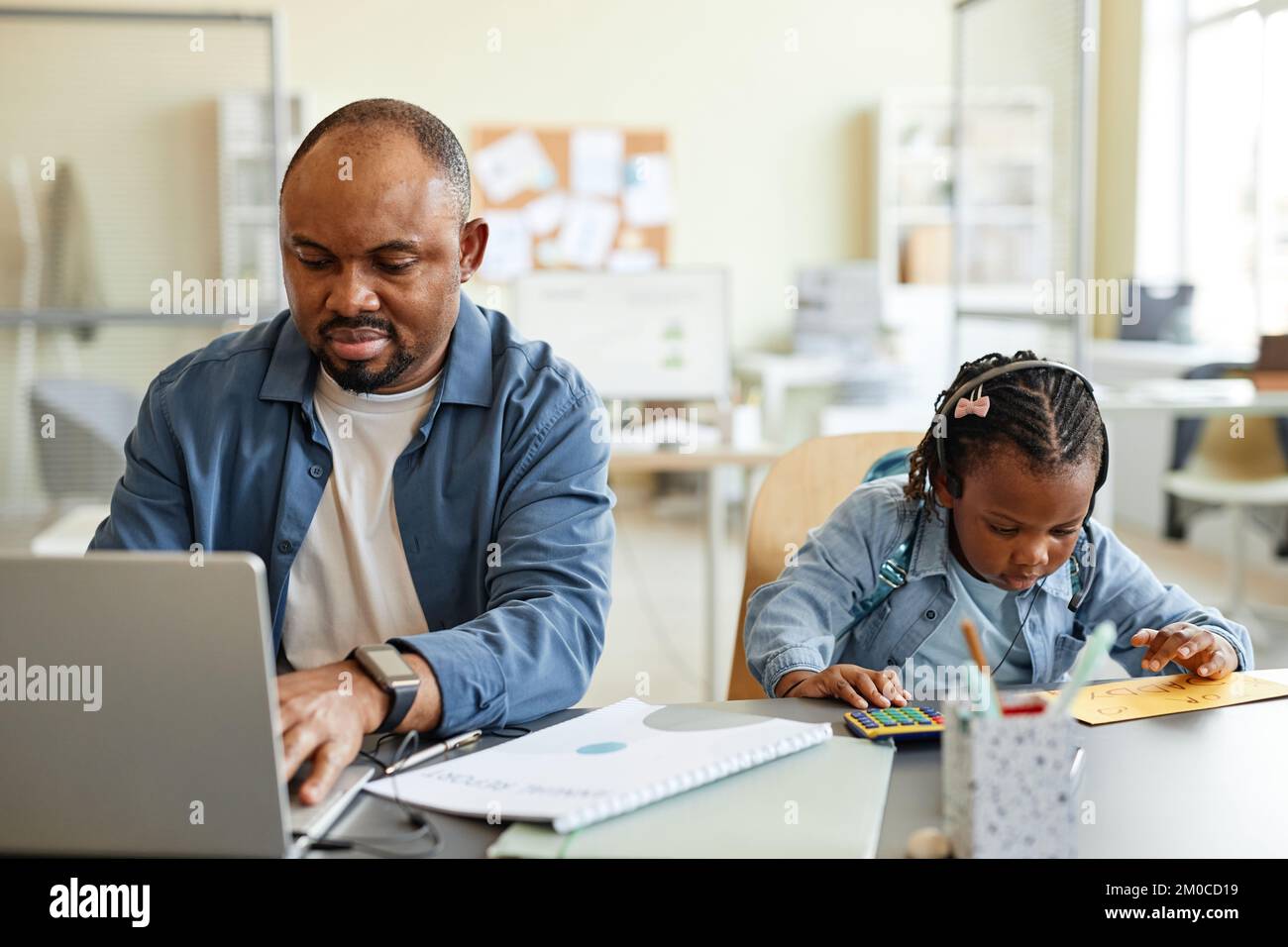 Vorderansicht eines schwarzen alleinerziehenden Vaters, der von zu Hause aus arbeitet, mit einem Kind, das mit dem Taschenrechner neben ihm spielt Stockfoto
