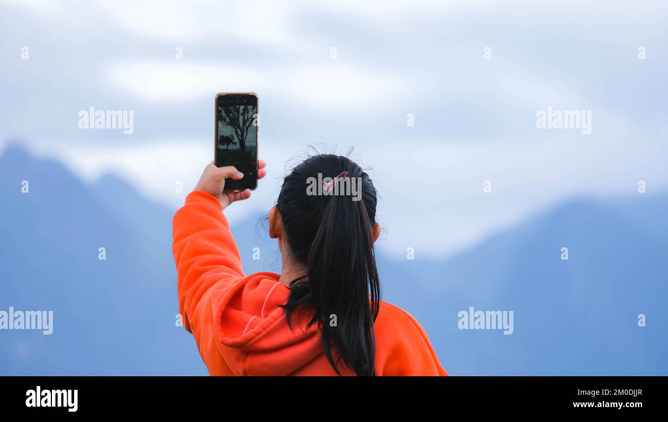 Rückansicht einer jungen Frau, die auf einem ruhigen Berg steht und am Telefon Fotos von der wunderschönen Berglandschaft bei Sonnenaufgang macht. Stockfoto