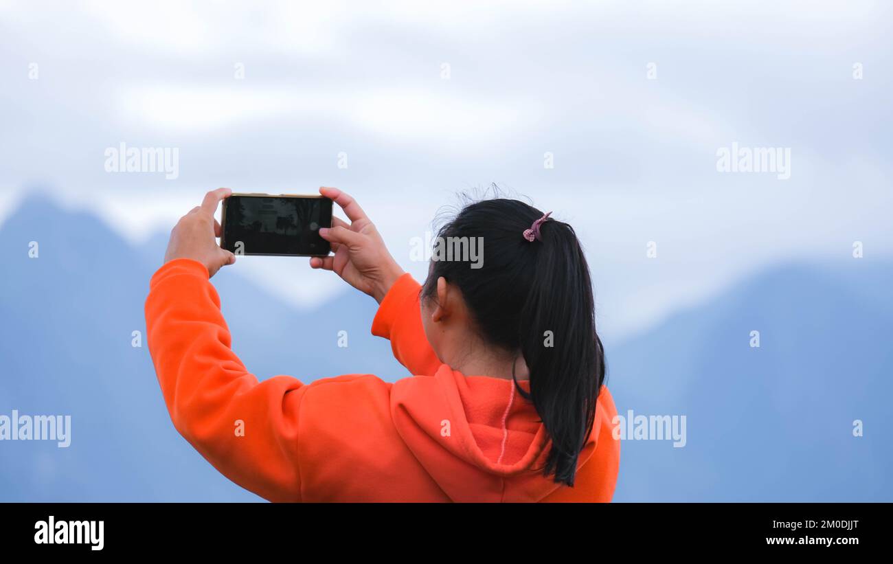 Rückansicht einer jungen Frau, die auf einem ruhigen Berg steht und am Telefon Fotos von der wunderschönen Berglandschaft bei Sonnenaufgang macht. Stockfoto